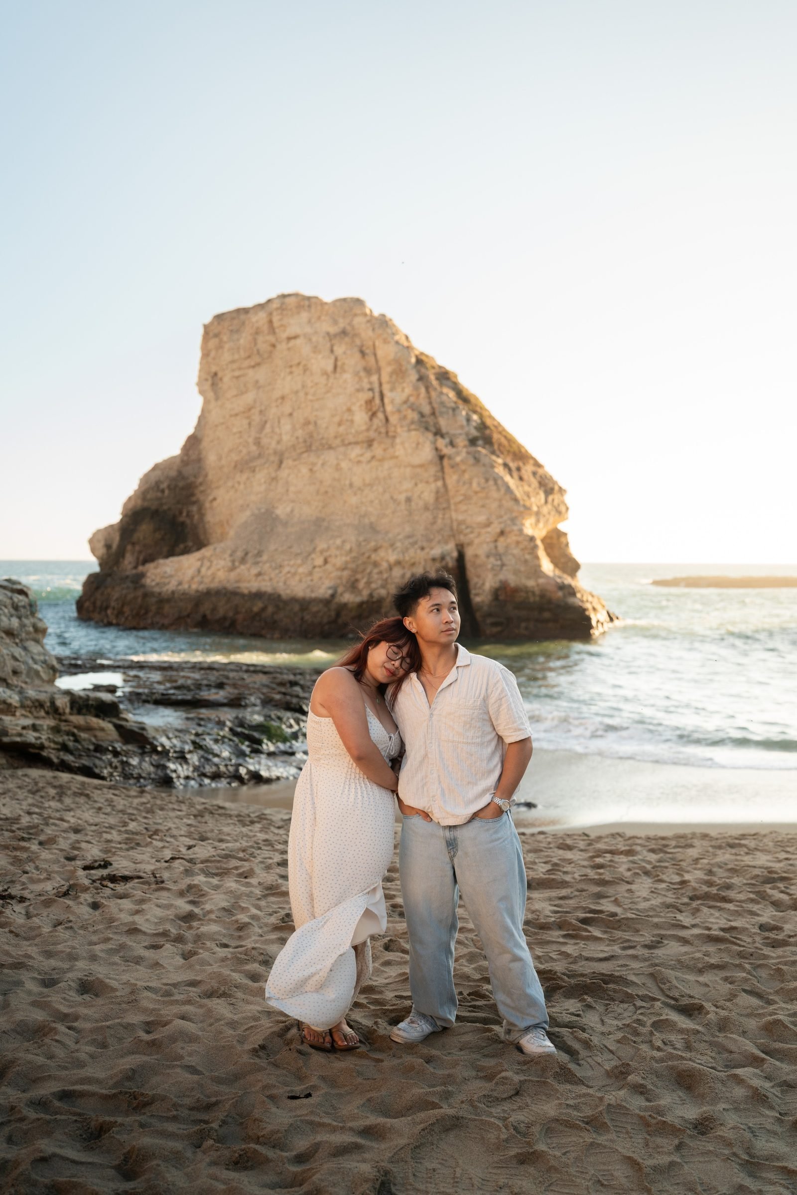 A couple stands on the sandy beach at Shark Fin Cove during golden hour. She leans her head on his shoulder in a white maxi dress while he gazes upward in a light button-down and pale jeans, a dramatic sea rock glowing behind them.