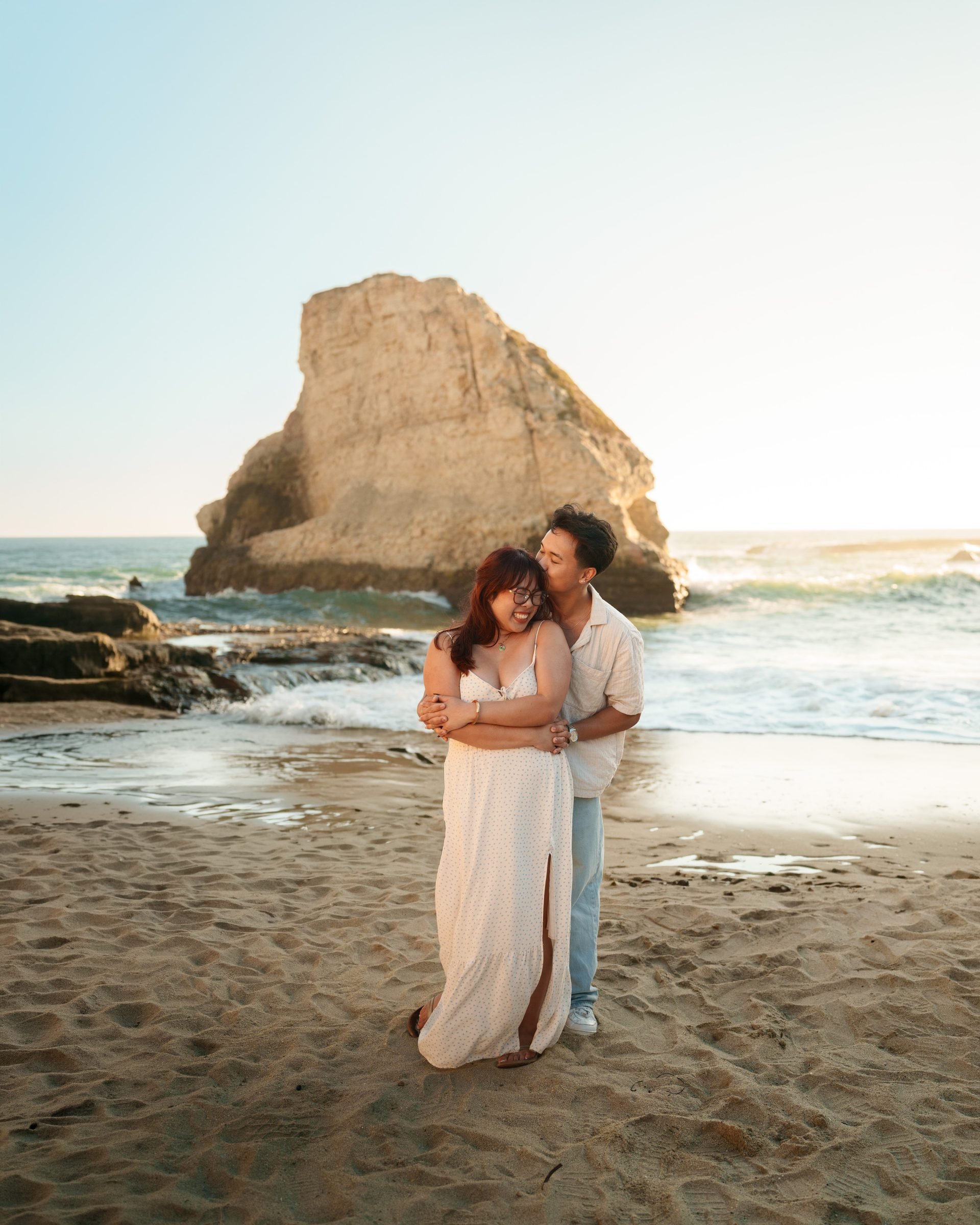 A couple embraces on the sand at Shark Fin Cove during golden hour, the iconic sea stack glowing warm behind them. She wears a white floral maxi dress; he wears a cream shirt and light jeans.