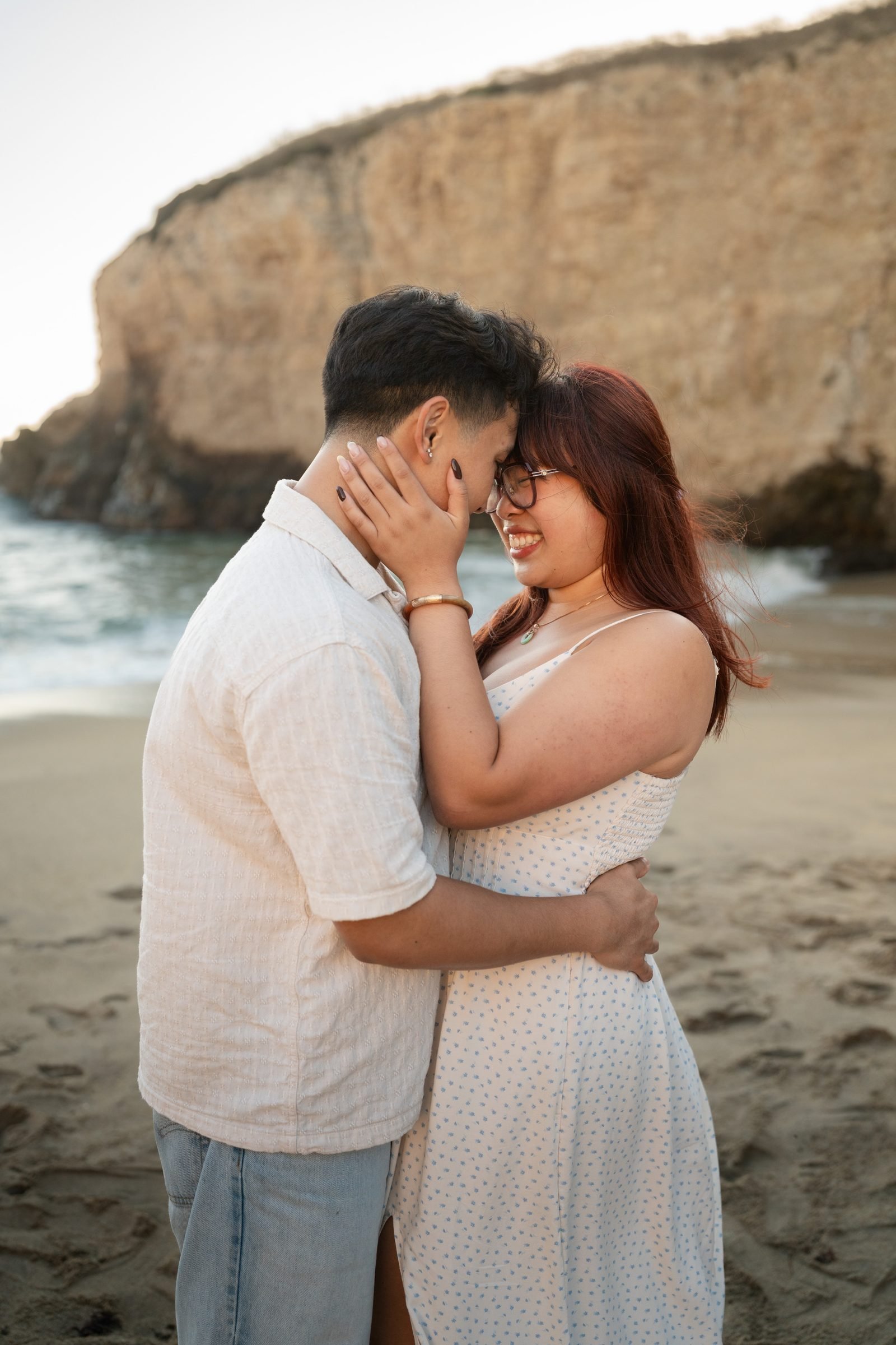 Couple sharing a close forehead-to-forehead moment on the sandy beach at Shark Fin Cove during golden hour, she laughs warmly with her hand cradling his face, both in neutral earth-tone outfits with a dramatic sandstone cliff behind them.