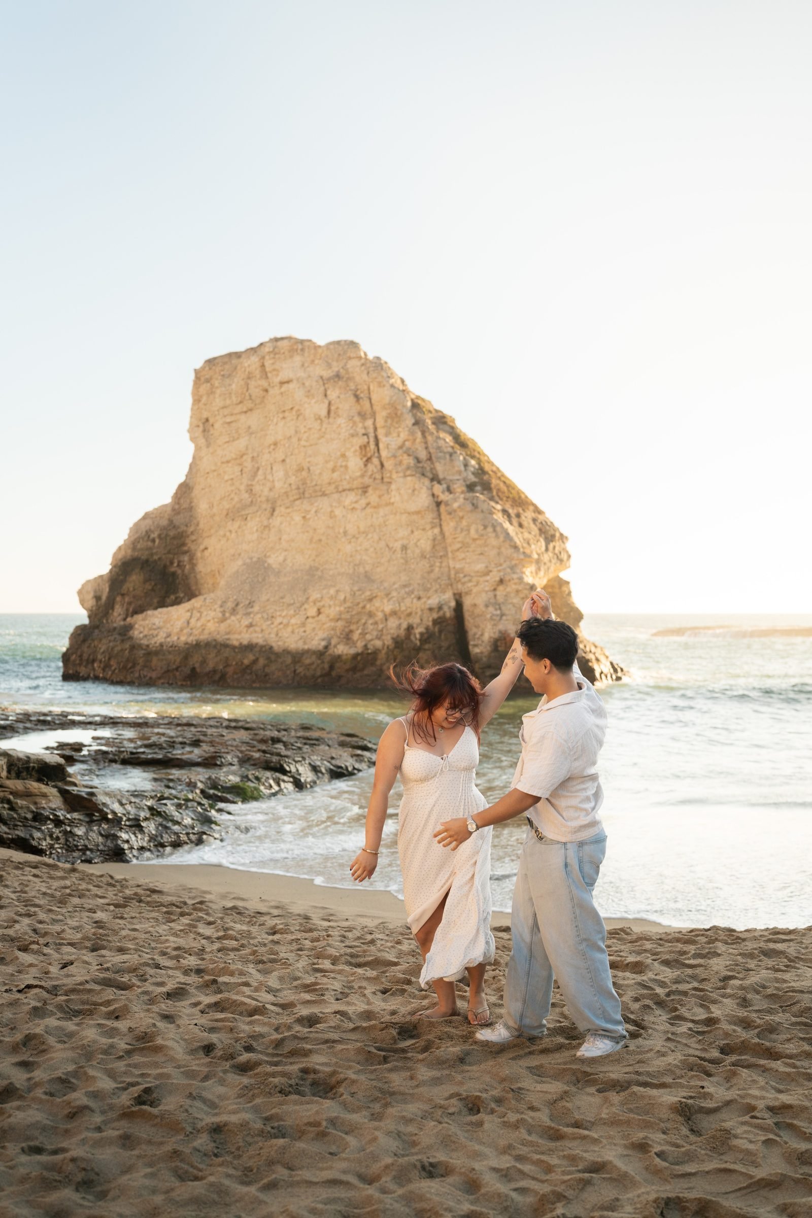 A couple dances and spins on the sandy shore at Shark Fin Cove during golden hour, the iconic rock formation glowing warm amber behind them as ocean waves roll in softly.