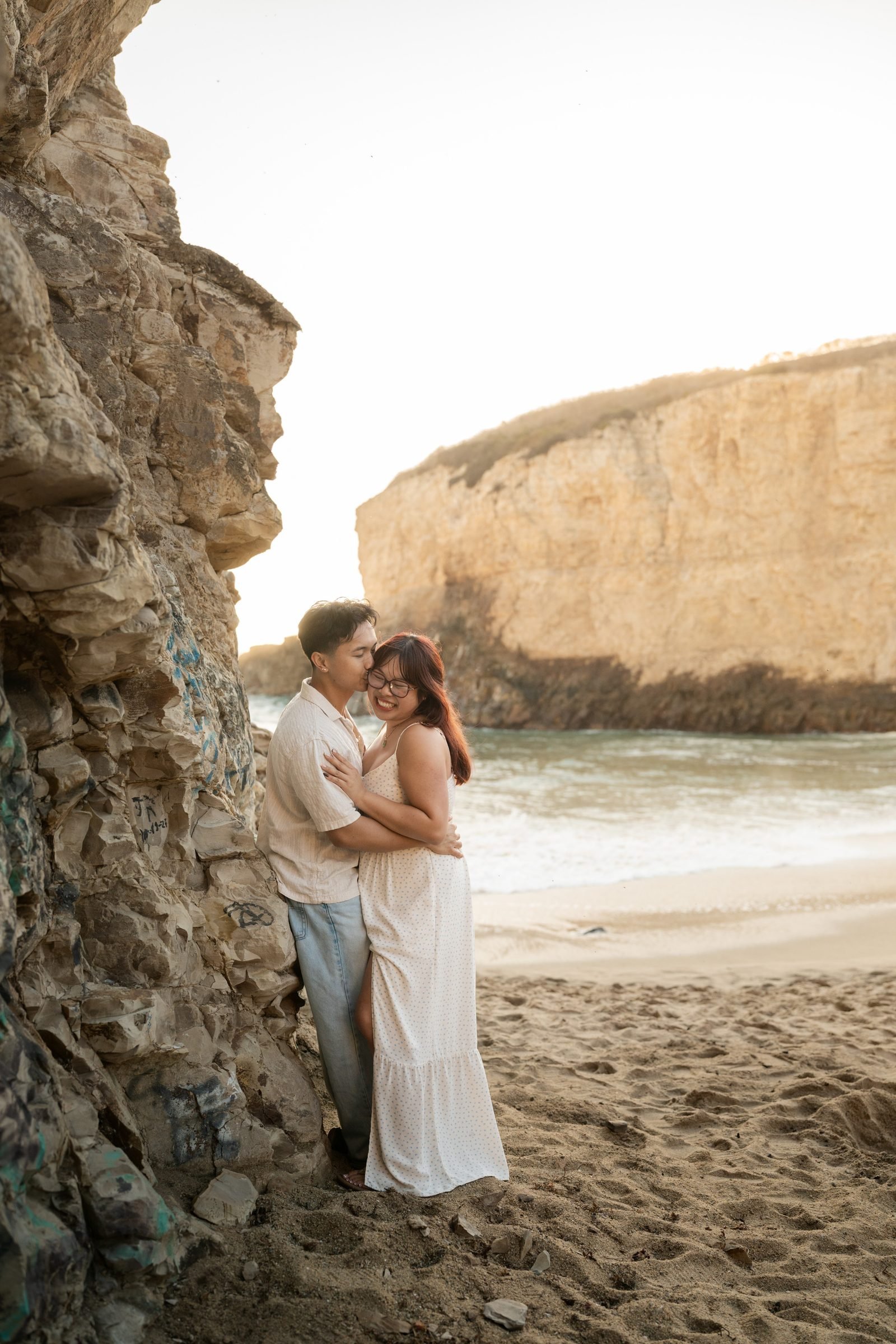 Couple leaning against a textured sea cliff at Shark Fin Cove during golden hour, he kisses her temple as she laughs in a white floral maxi dress, warm sandstone bluff visible in the background.