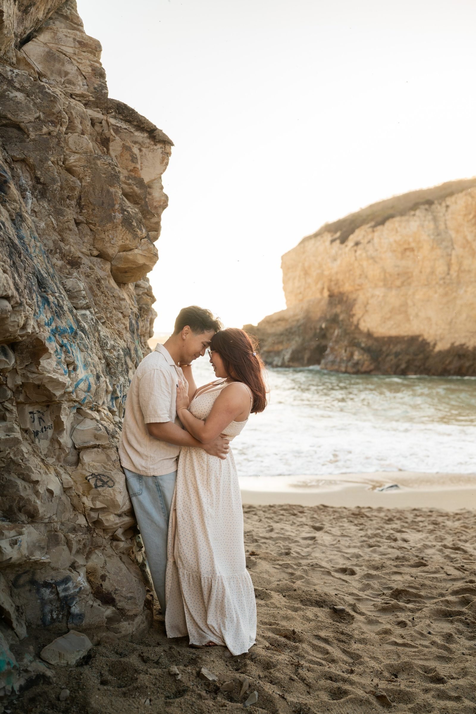 Couple leaning foreheads together against a weathered sandstone cliff at Shark Fin Cove, bathed in warm golden hour light, with the Pacific shoreline and the iconic fin-shaped rock glowing in the background.