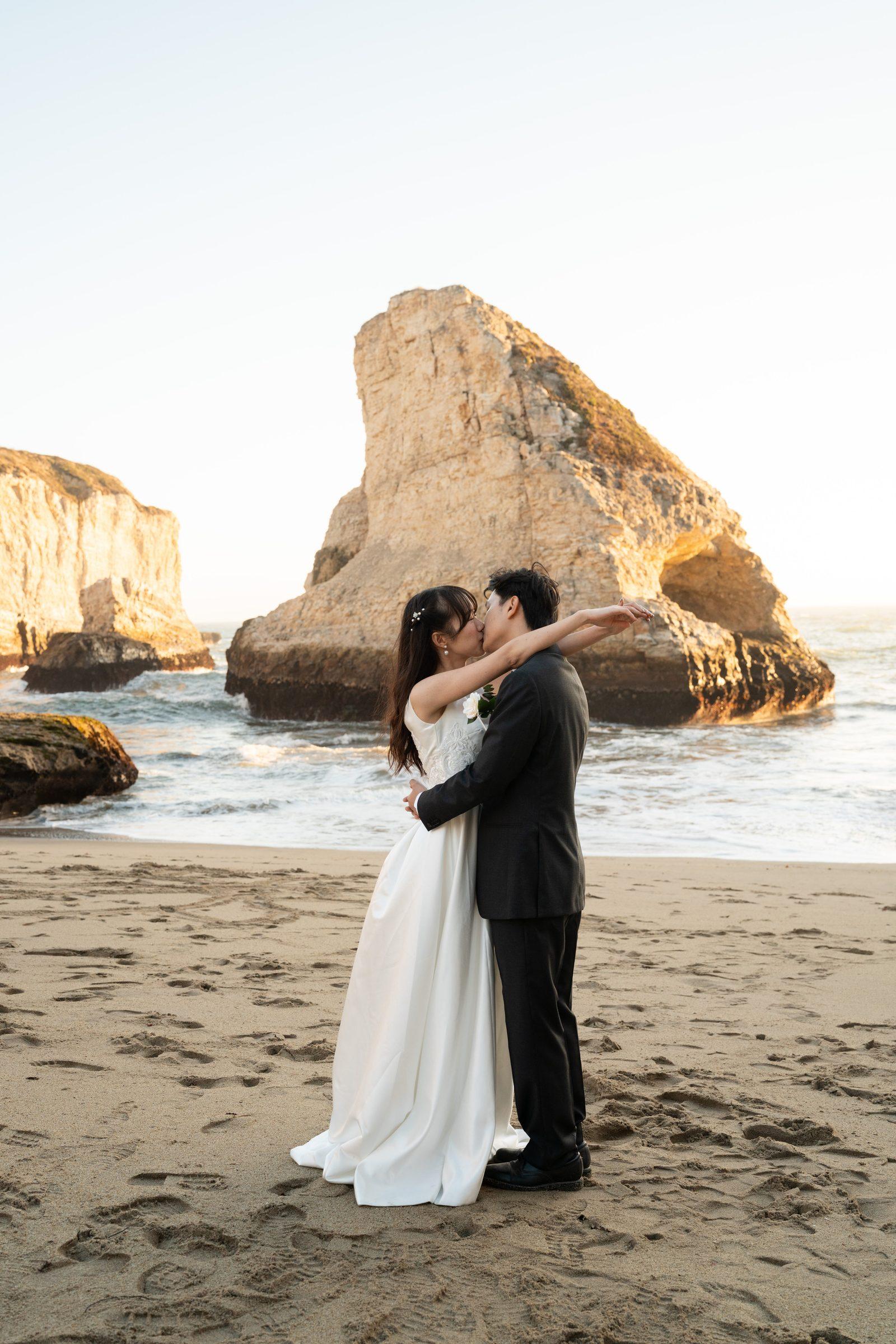 A couple shares a kiss on the sandy beach at Shark Fin Cove during golden hour, framed by dramatic sea stacks and ocean waves. She wears a flowing white gown, he wears a dark suit.