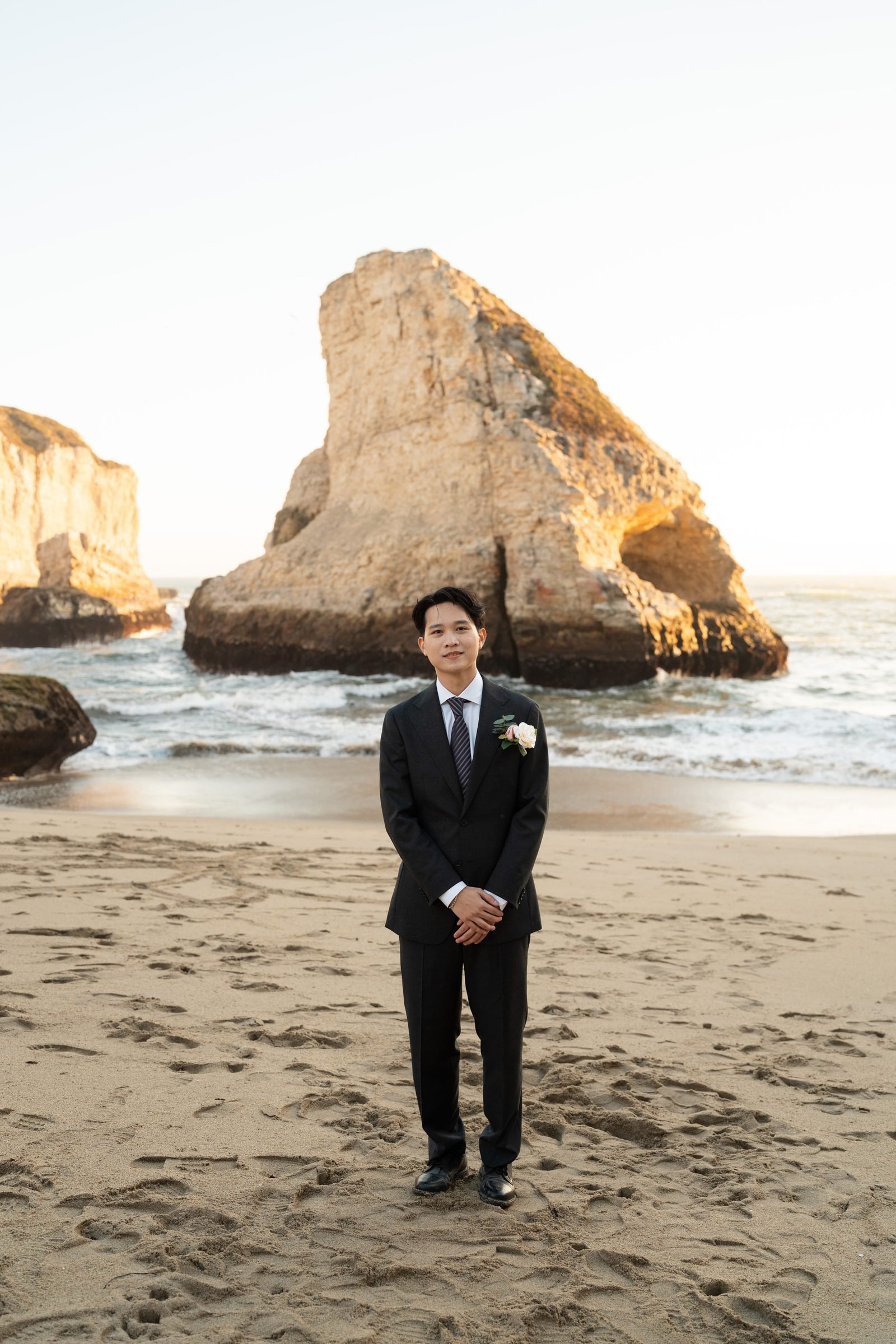 Groom in a dark suit with cream rose boutonniere stands on Shark Fin Cove beach at golden hour, dramatic sea stack glowing amber behind him, hands clasped in front