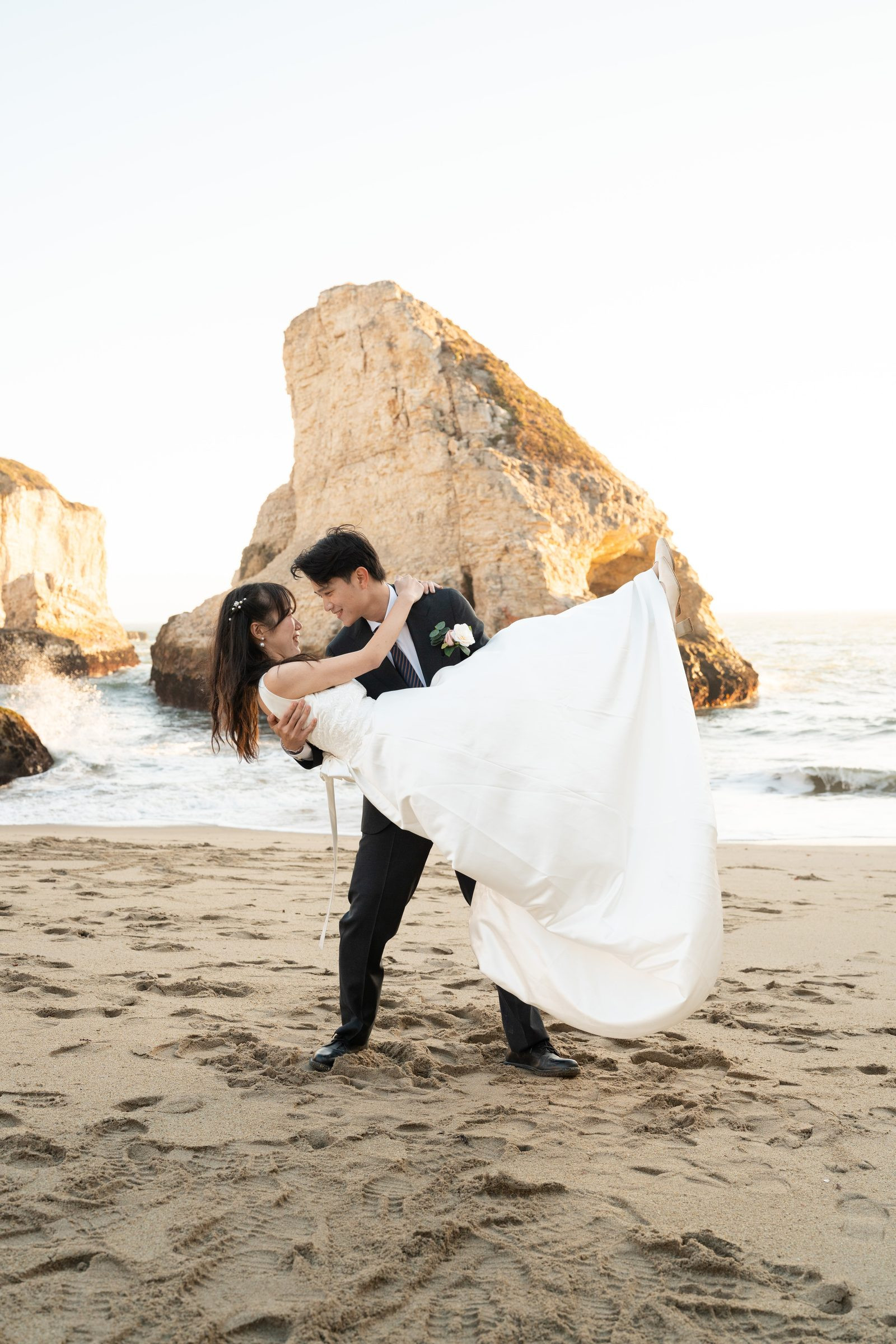 Groom in a dark suit lifts his bride in a flowing white gown on the sandy beach at Shark Fin Cove, golden hour light warming the dramatic sea stacks and crashing waves behind them.