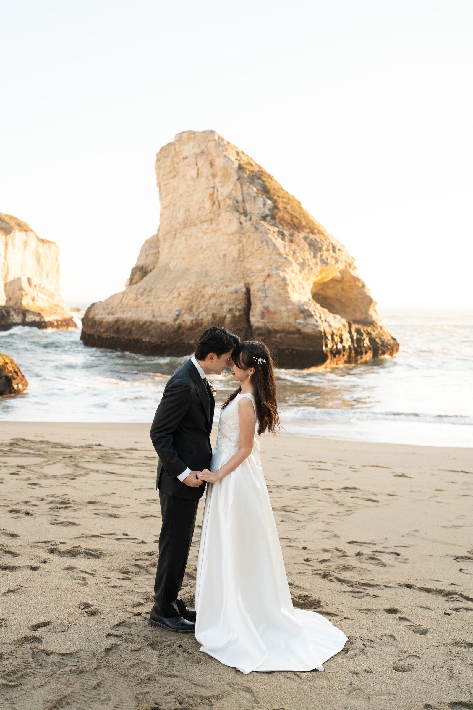 A couple in wedding attire stand forehead-to-forehead on the sand at Shark Fin Cove, holding hands, with a dramatic sea stack and golden-hour ocean light glowing behind them.