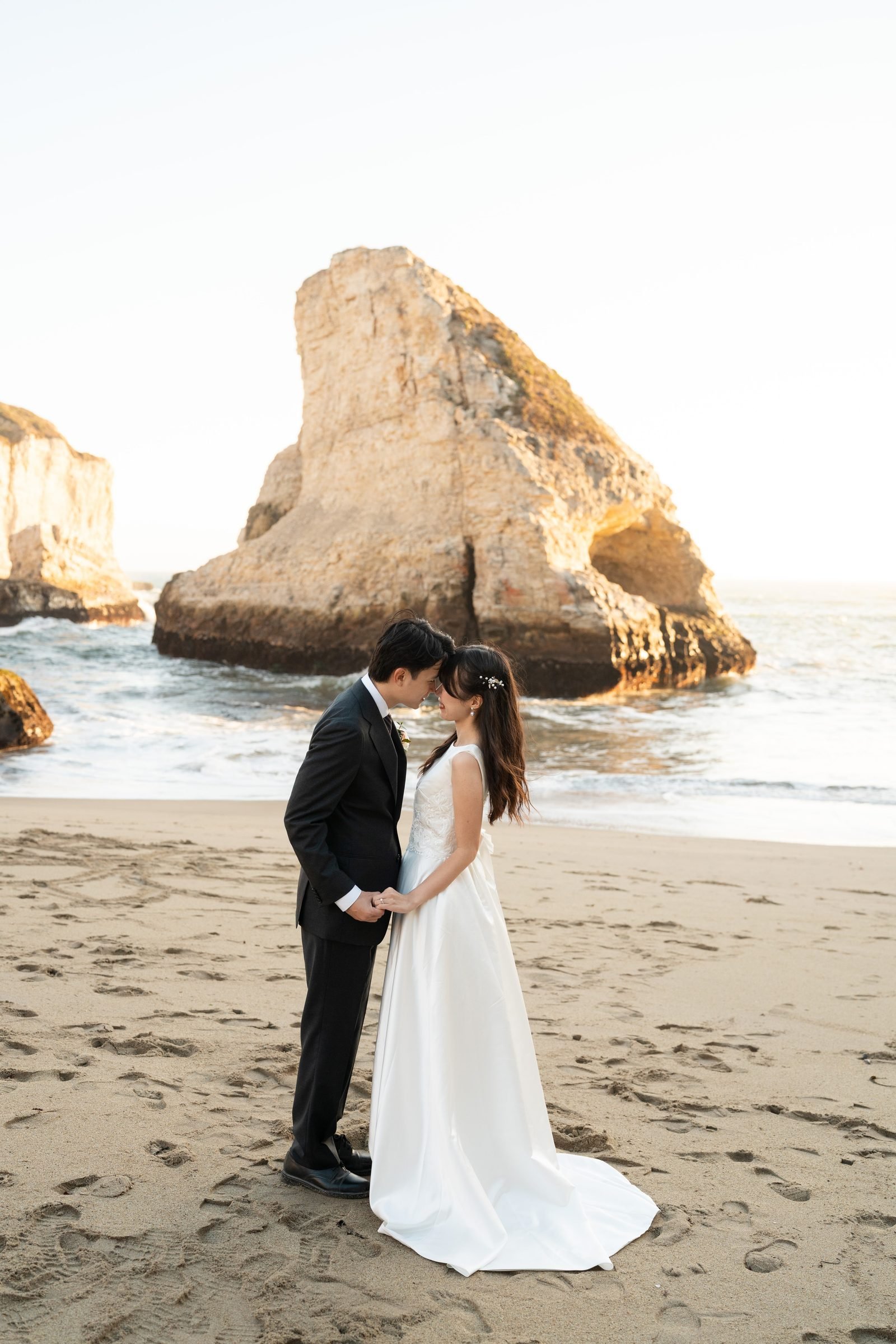 A couple in wedding attire stand forehead-to-forehead on the sand at Shark Fin Cove, holding hands, with a dramatic sea stack and golden-hour ocean light glowing behind them.