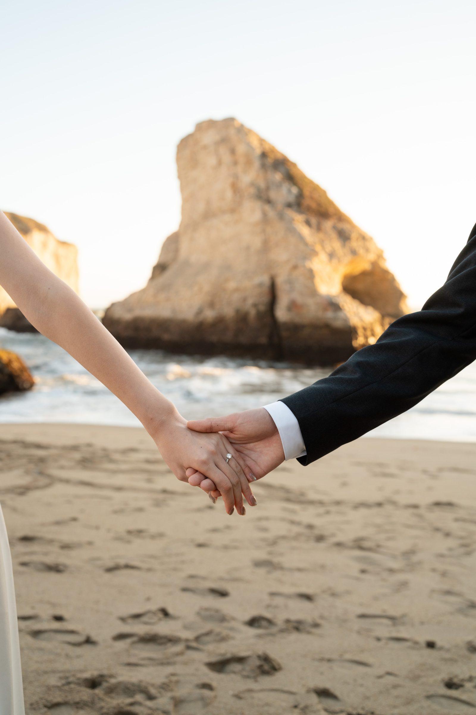 Close-up of a couple holding hands on a sandy beach at golden hour, with a dramatic sea stack rising from the Pacific behind them. Her engagement ring catches the warm light; he wears a dark suit jacket.