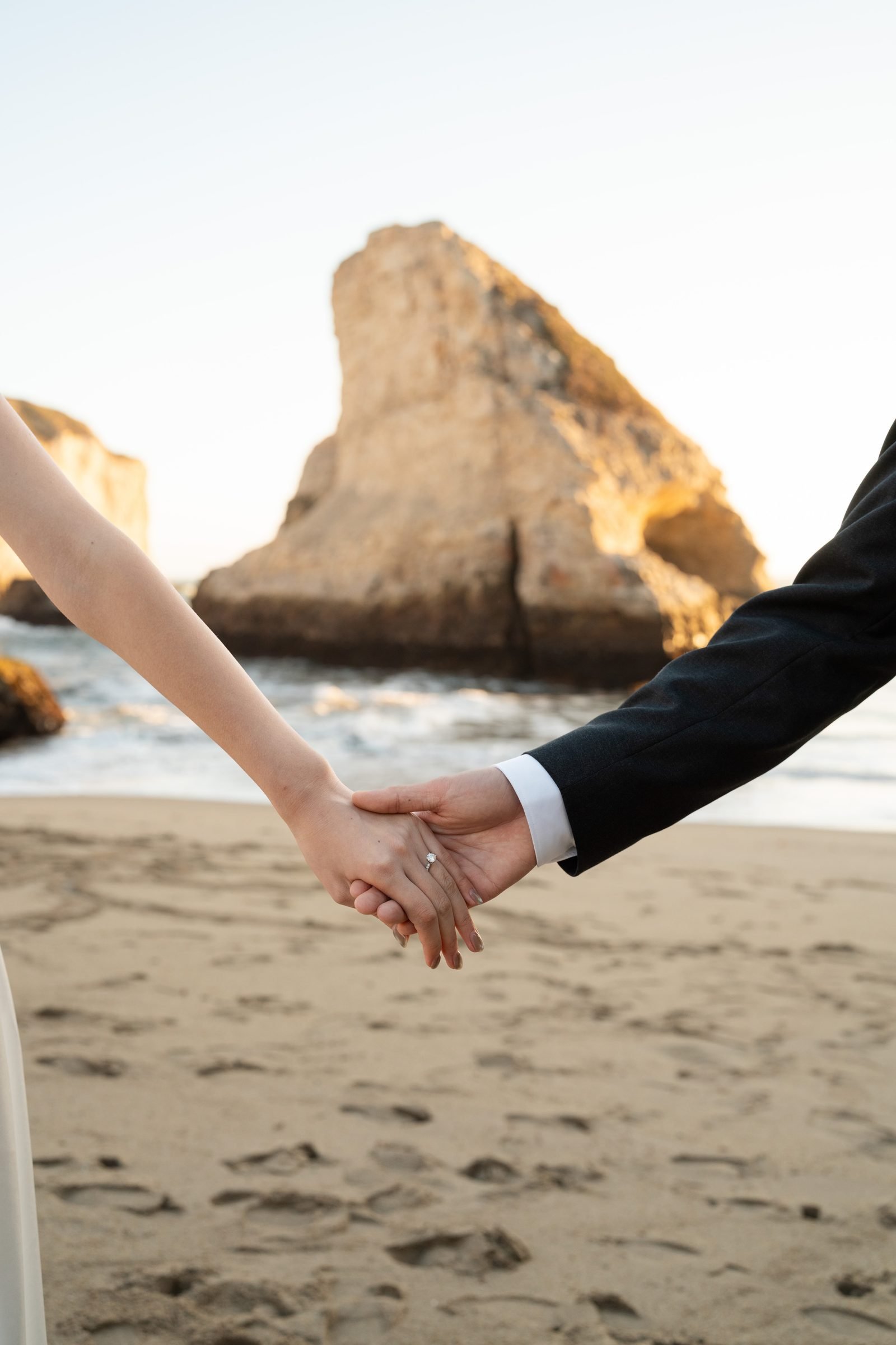 Close-up of a couple holding hands on a sandy beach at golden hour, with a dramatic sea stack rising from the Pacific behind them. Her engagement ring catches the warm light; he wears a dark suit jacket.