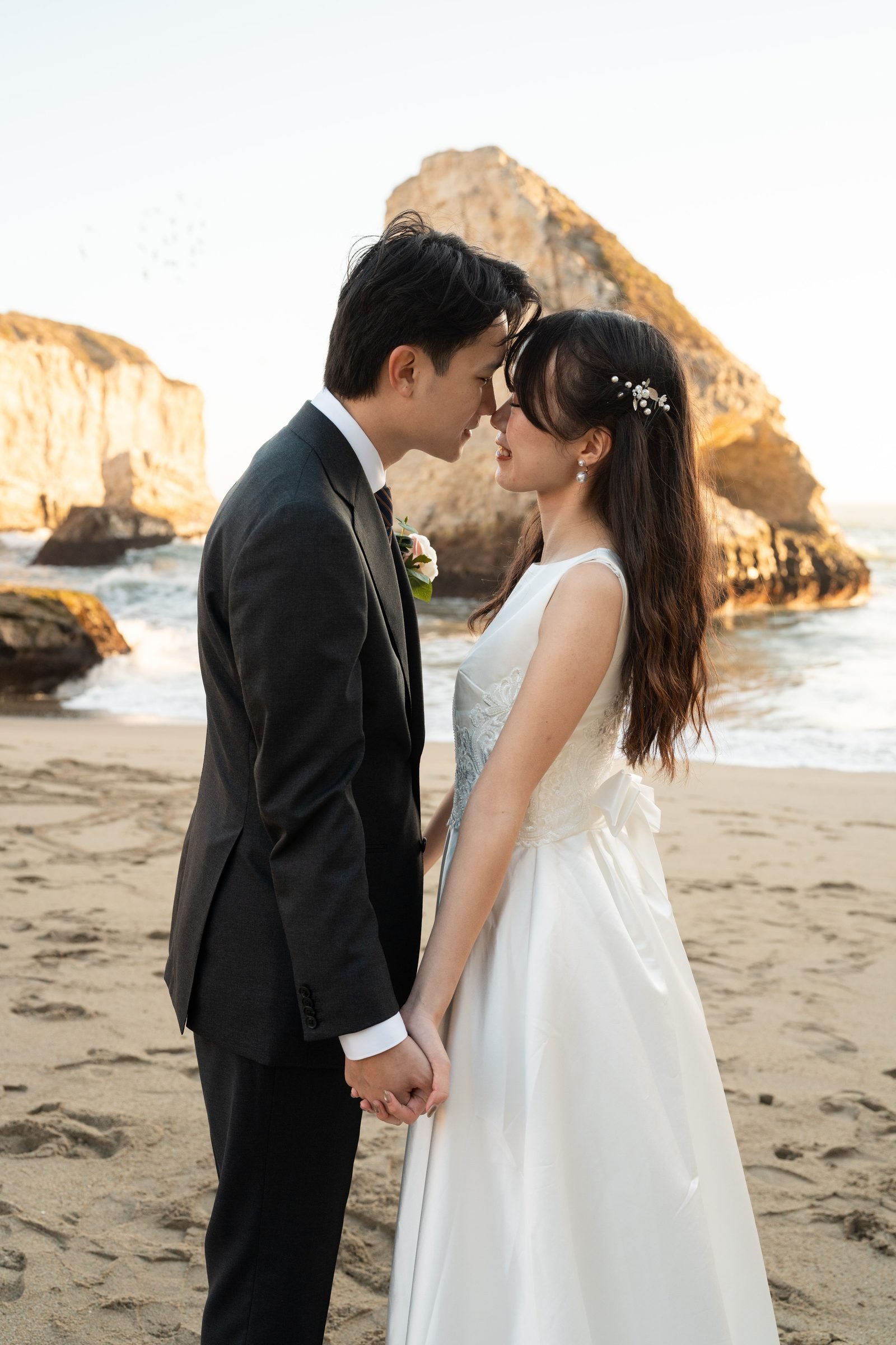 Couple in wedding attire touching foreheads on the sand at Shark Fin Cove during golden hour, with dramatic sea stacks and ocean waves behind them.