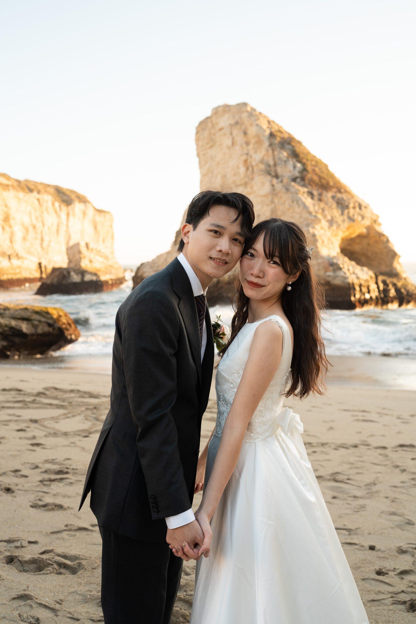 A couple stands hand-in-hand on the sandy beach at Shark Fin Cove during golden hour, cheeks pressed together as they smile softly at the camera, with dramatic sea stacks glowing amber behind them.
