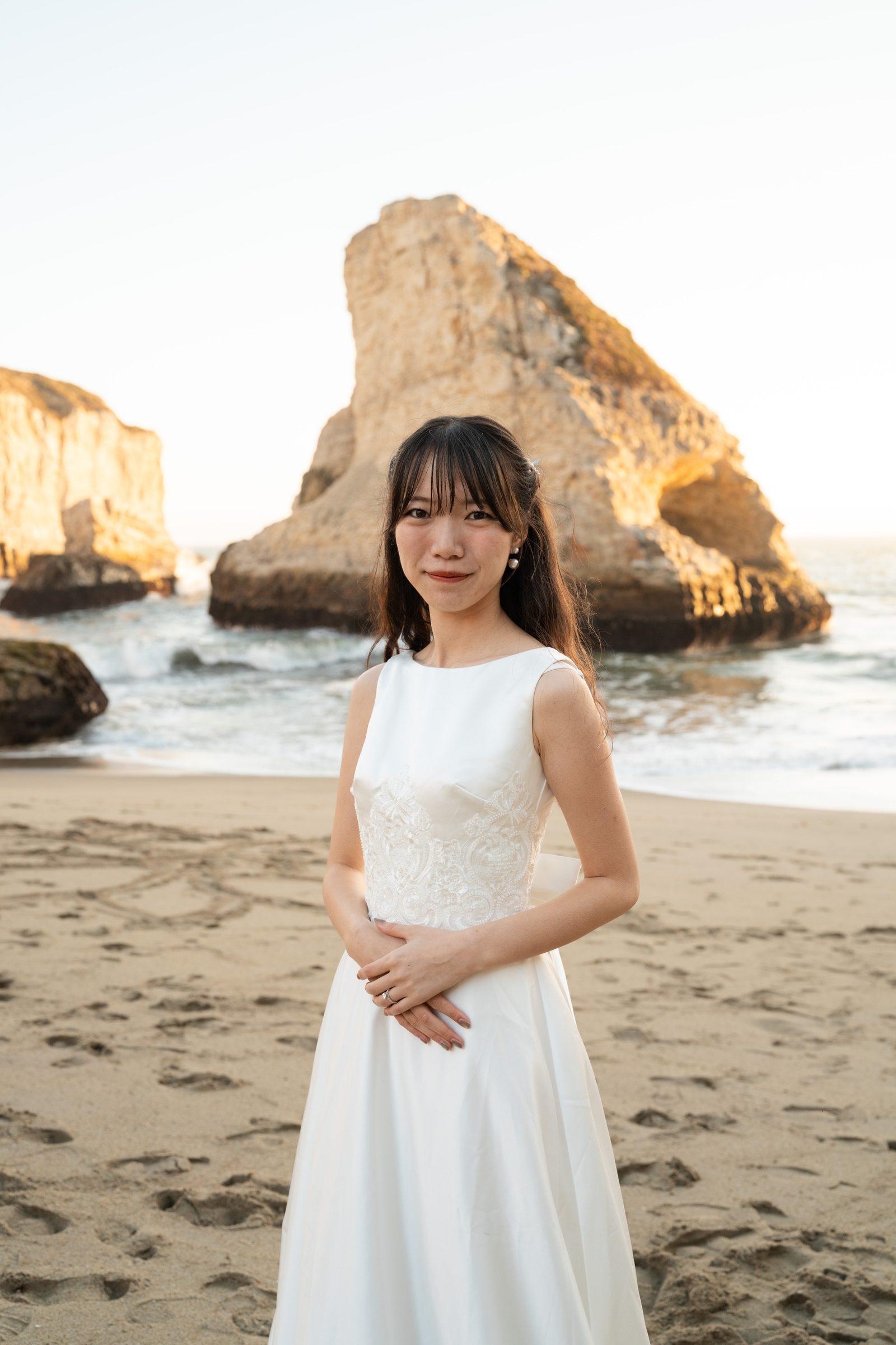 Bride in a sleeveless white gown with lace bodice stands on Shark Fin Cove beach at golden hour, dramatic sea stack glowing amber behind her, hands gently clasped at her waist.