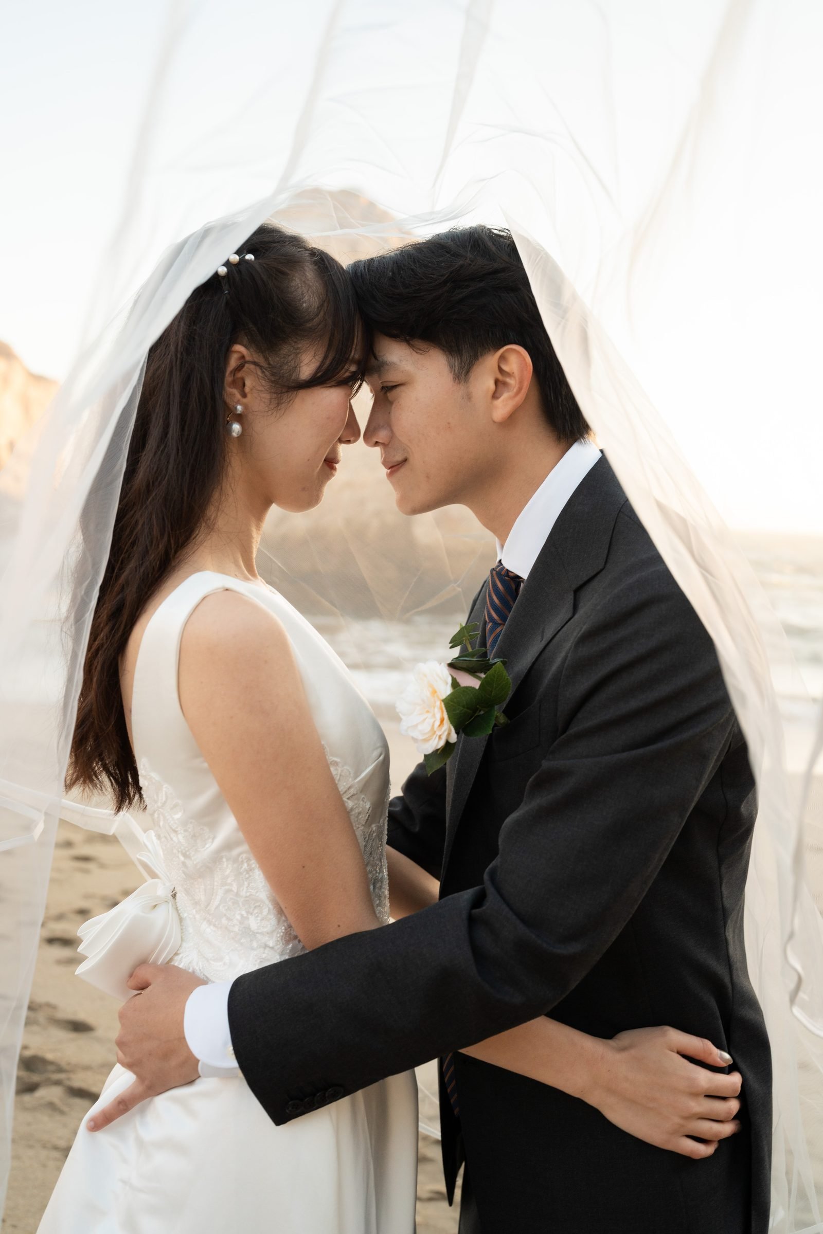 Couple pressed forehead to forehead under a billowing white veil on Shark Fin Cove beach at golden hour, his arms wrapped around her waist, both smiling softly with eyes closed