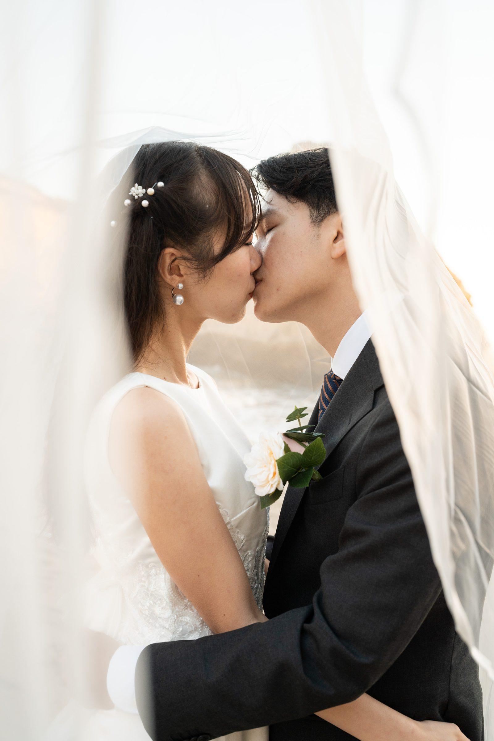 Couple sharing a tender kiss under a flowing white veil, her pearl hair pins and drop earrings catching soft diffused light, his dark suit and cream boutonniere pressed close against her white lace-bodice gown.