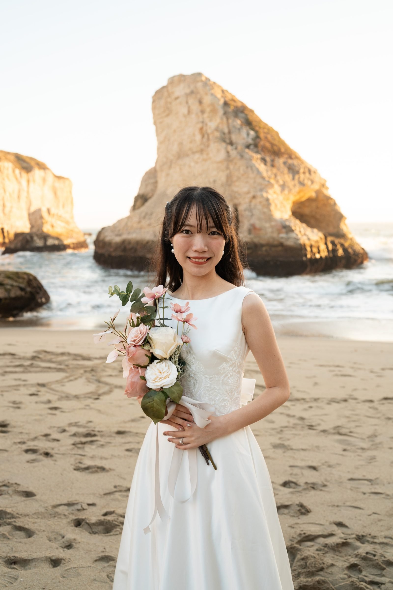 Woman in a sleeveless white bridal gown holding a blush and ivory bouquet stands on sandy beach at Shark Fin Cove during golden hour, dramatic sea stacks rising from the ocean behind her