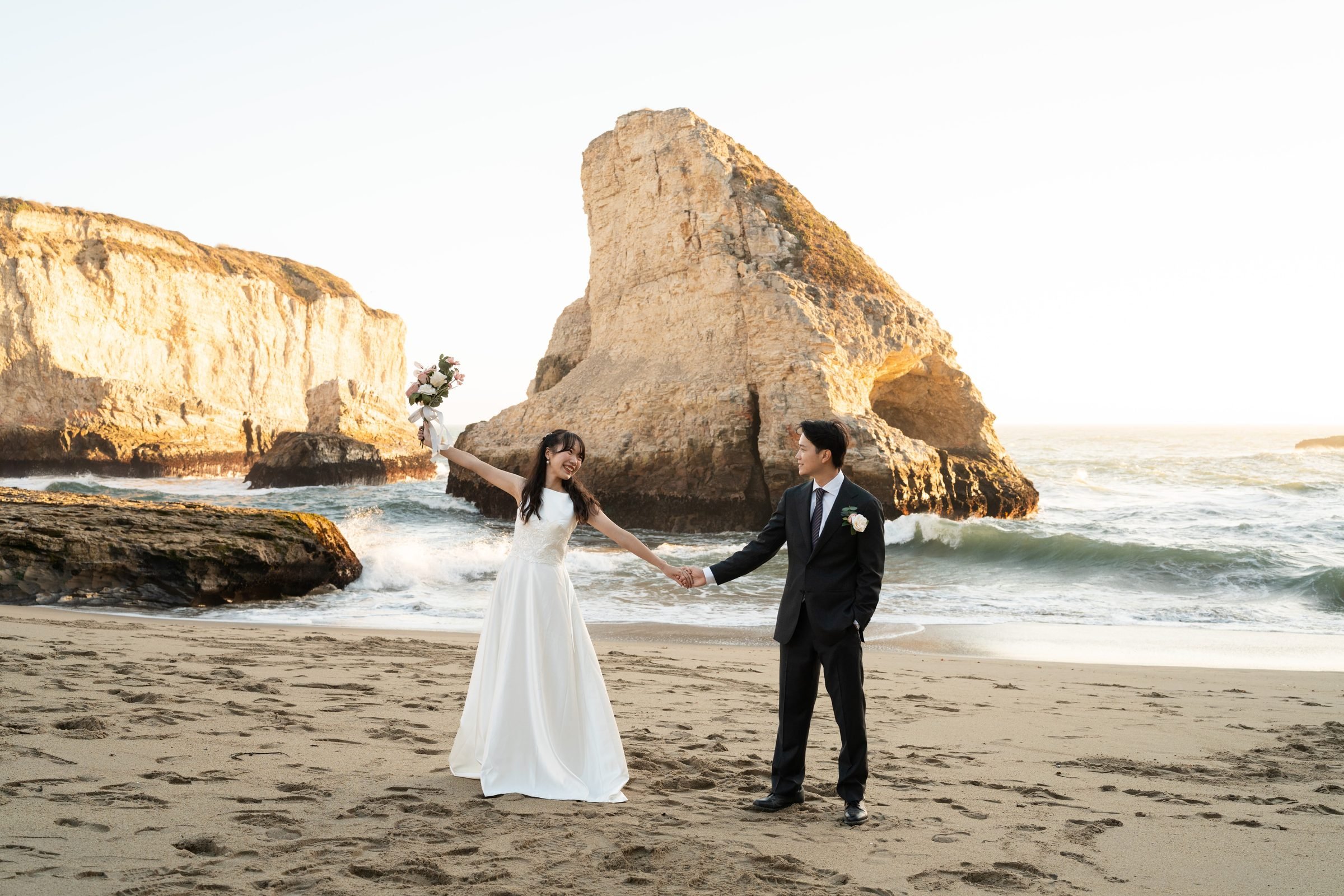Couple in wedding attire holding hands on the sandy beach at Shark Fin Cove during golden hour, bride raising bouquet overhead with a joyful laugh as dramatic sea stacks and crashing waves fill the background.