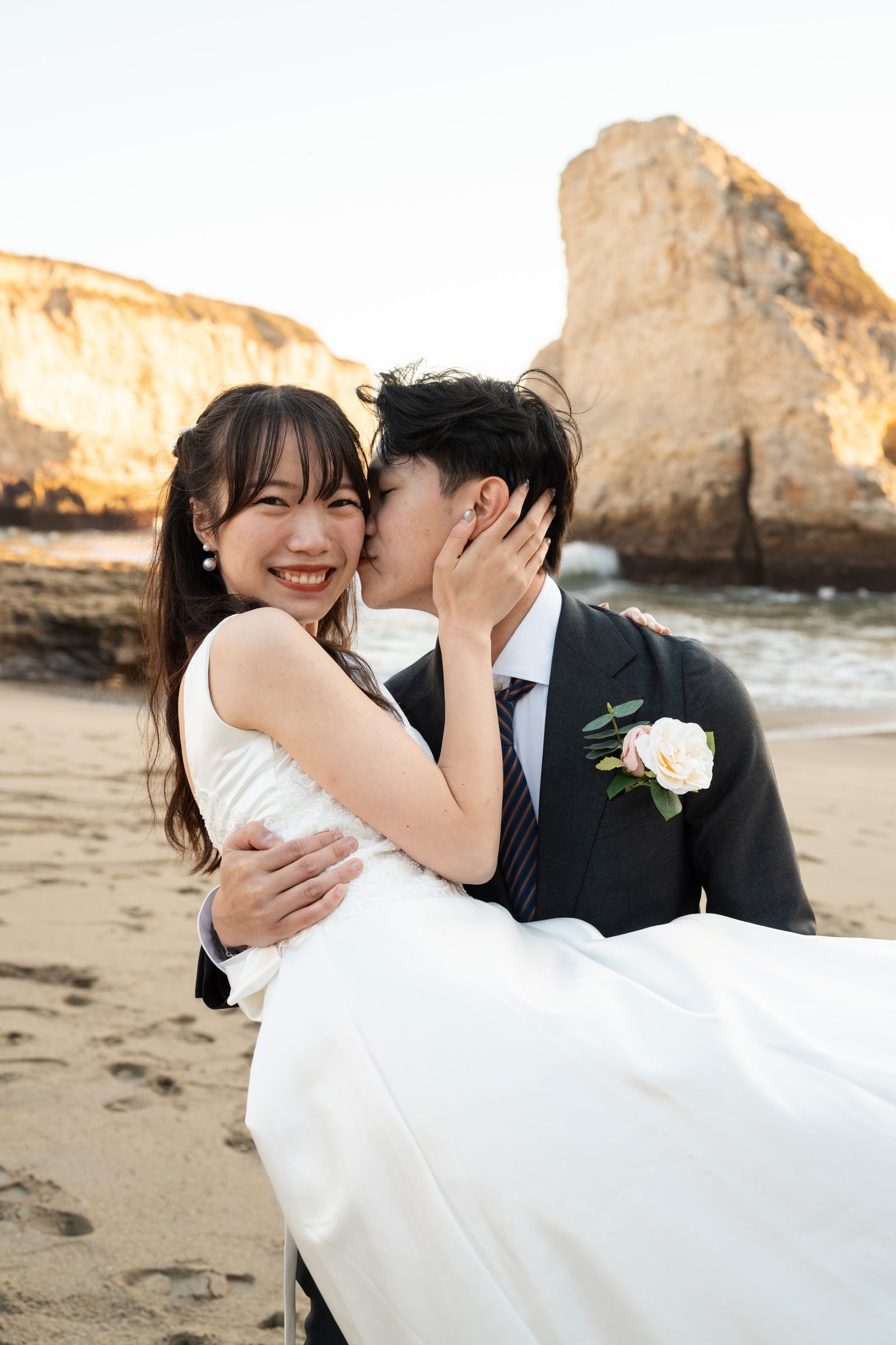 A man in a dark suit with a blush boutonnière carries a woman in a white gown on a sandy beach at golden hour, kissing her cheek as she laughs toward the camera with sea cliffs glowing behind them.