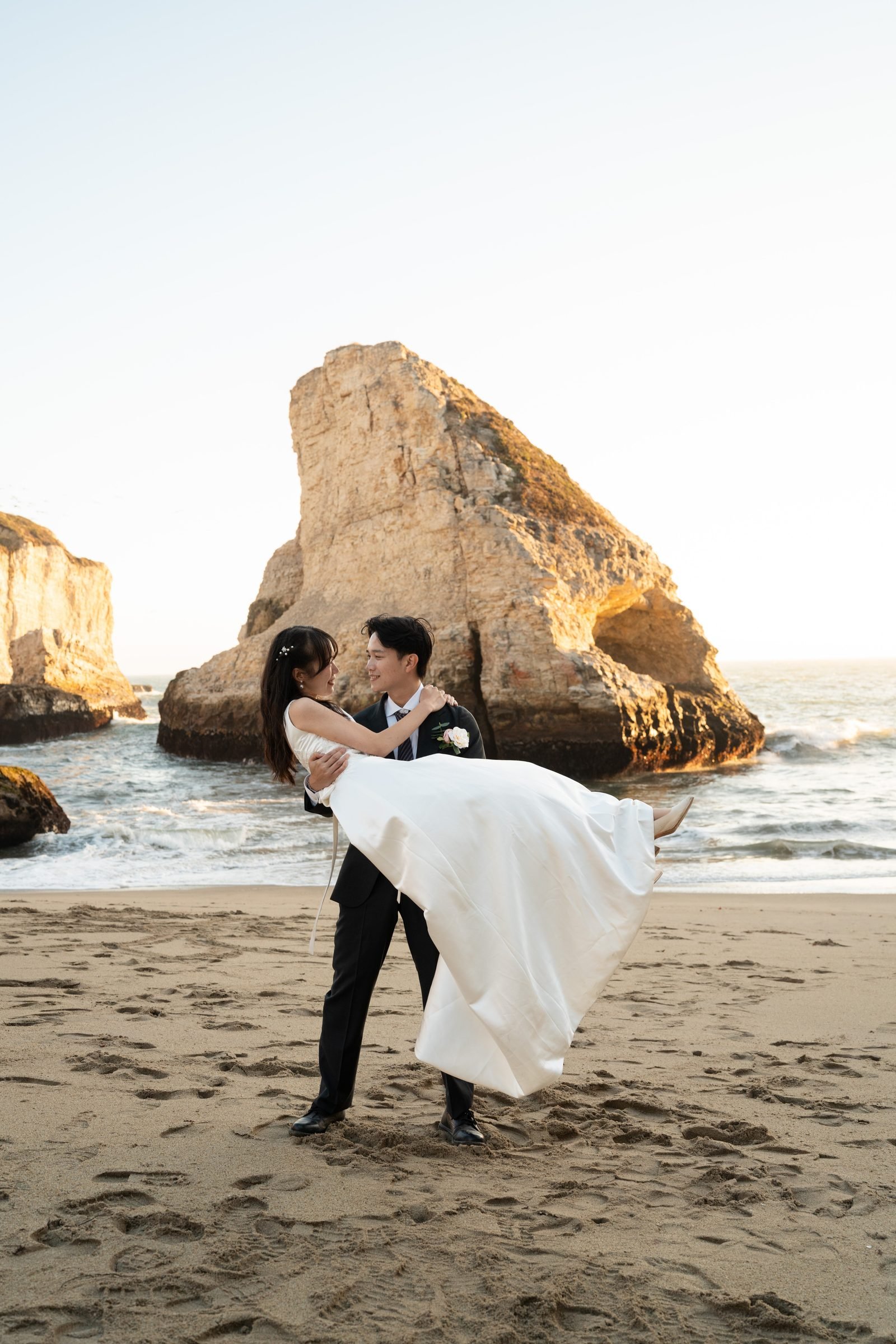 Groom in a black suit lifts his bride in a flowing white gown on the sand at Shark Fin Cove, both laughing and gazing at each other, with the iconic sea stack glowing in warm golden hour light behind them.