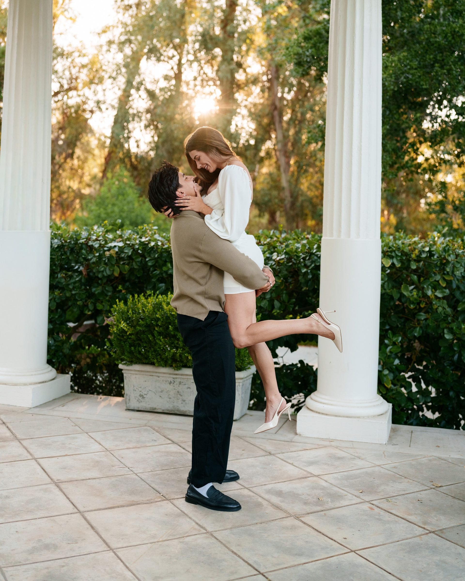 Man lifts woman between white columns on a stone terrace at Montalvo Arts Center during golden hour; she smiles down at him while he looks up at her, her slingback heels visible as the sun streams through the trees behind them.