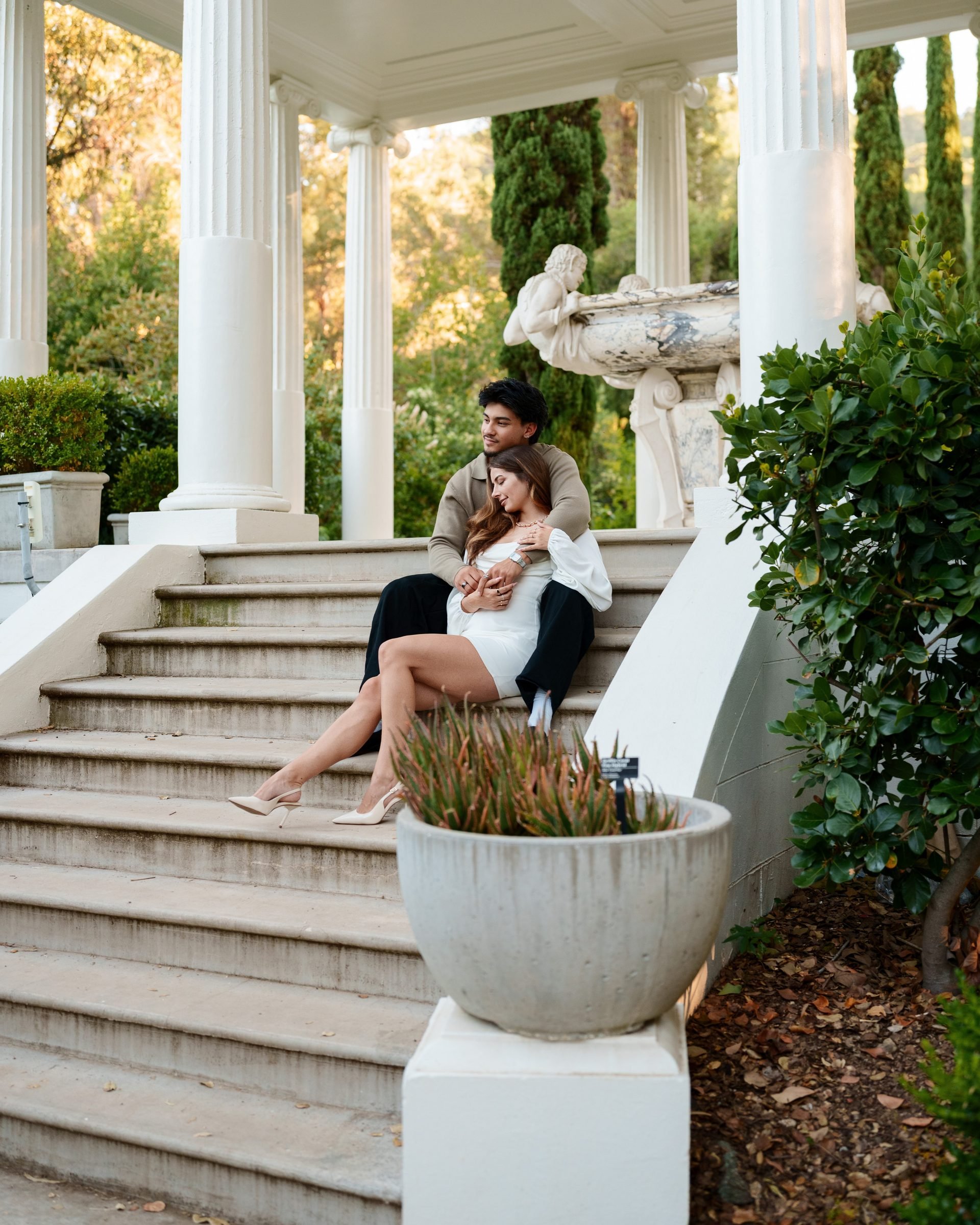 Couple sits nestled together on wide stone steps of a classical portico at Montalvo Arts Center at golden hour; she leans back into his arms with eyes closed while he gazes outward, a marble statue and cypress trees visible behind them.