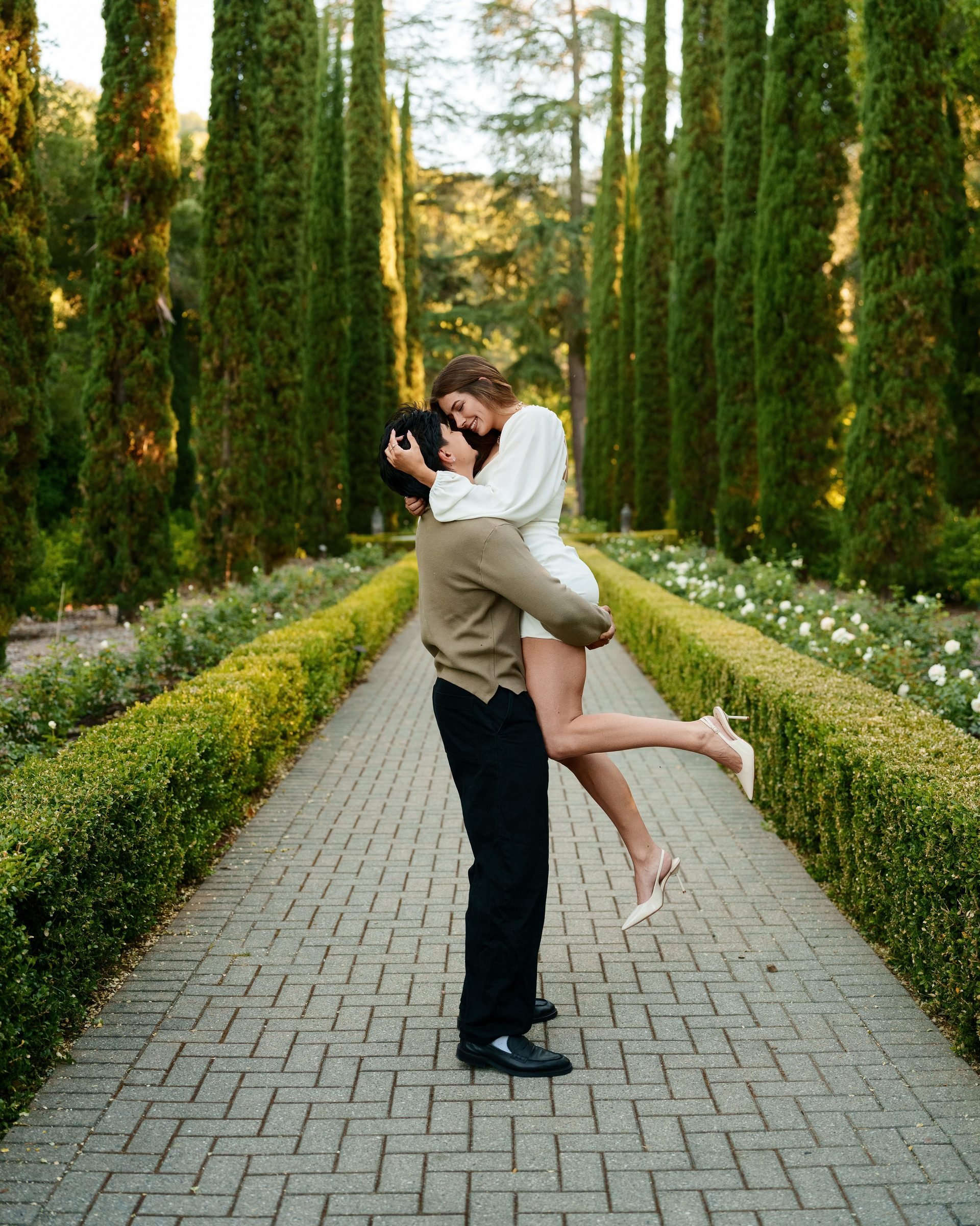 A man lifts his girlfriend off the ground at the center of Montalvo's cypress-lined garden allee. She wraps her arms around his neck and laughs as her leg kicks up. Golden hour light warms the lush green backdrop behind them.