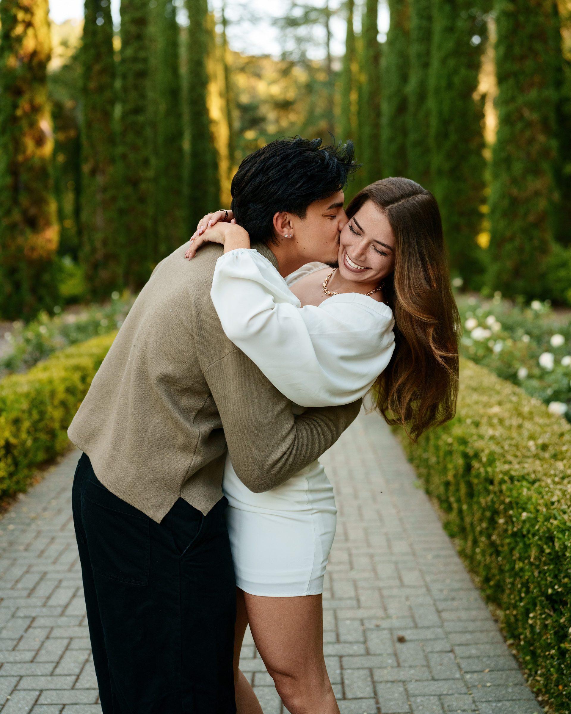 A young man plants a playful kiss on his laughing girlfriend's cheek along a formal garden path lined with tall Italian cypresses at golden hour. She wears a white mini dress; he wears a tan sweater and black trousers.