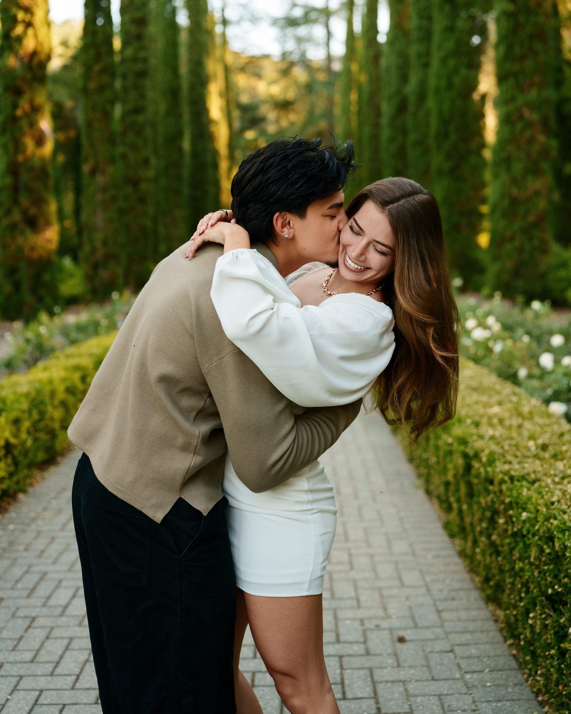 A young man plants a playful kiss on his laughing girlfriend's cheek along a formal garden path lined with tall Italian cypresses at golden hour. She wears a white mini dress; he wears a tan sweater and black trousers.