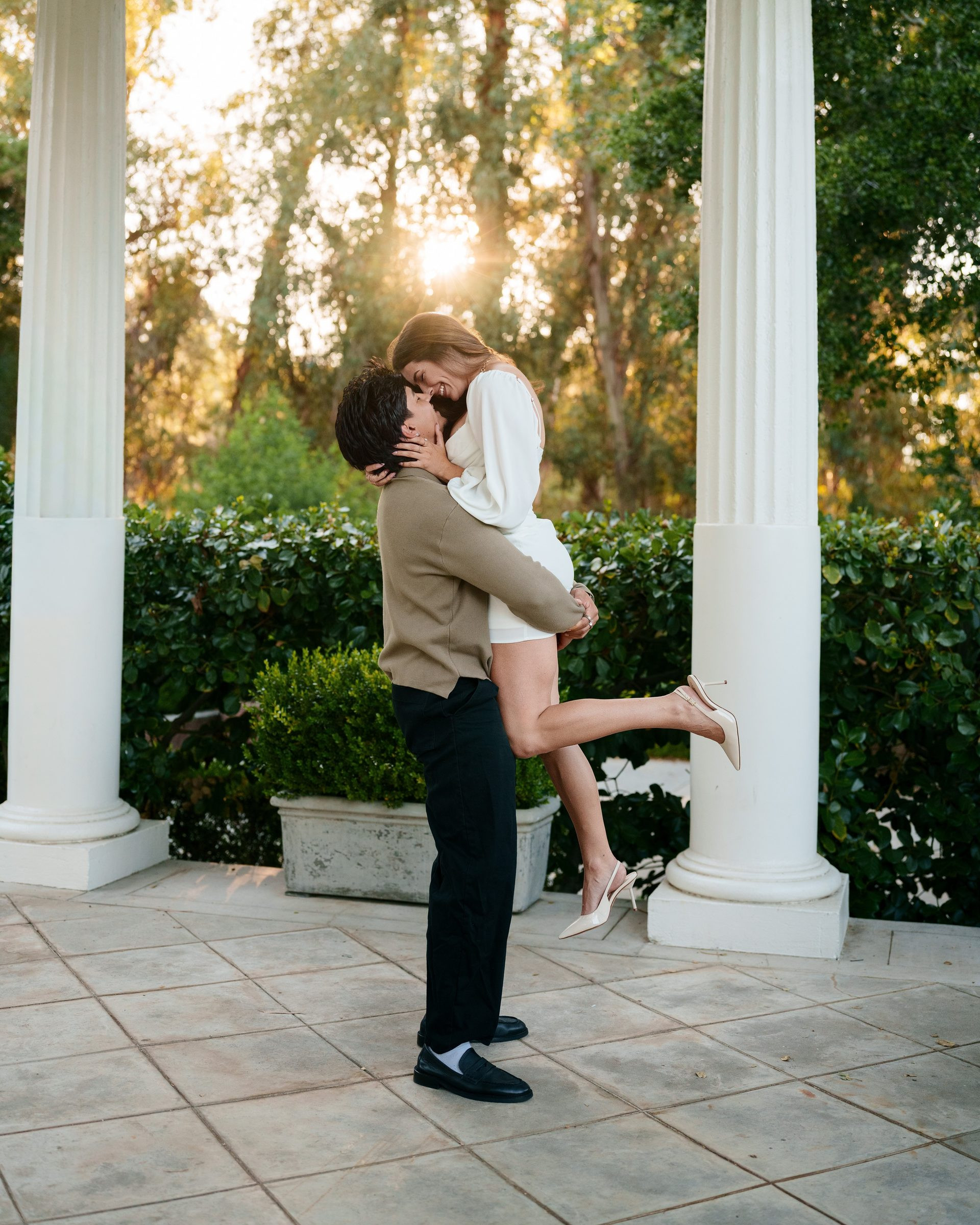 He lifts her off the ground for a kiss between white columns at Montalvo Arts Center at golden hour, sun bursting through the trees behind them, her ivory heels popped mid-air, both in a white dress and tan sweater.