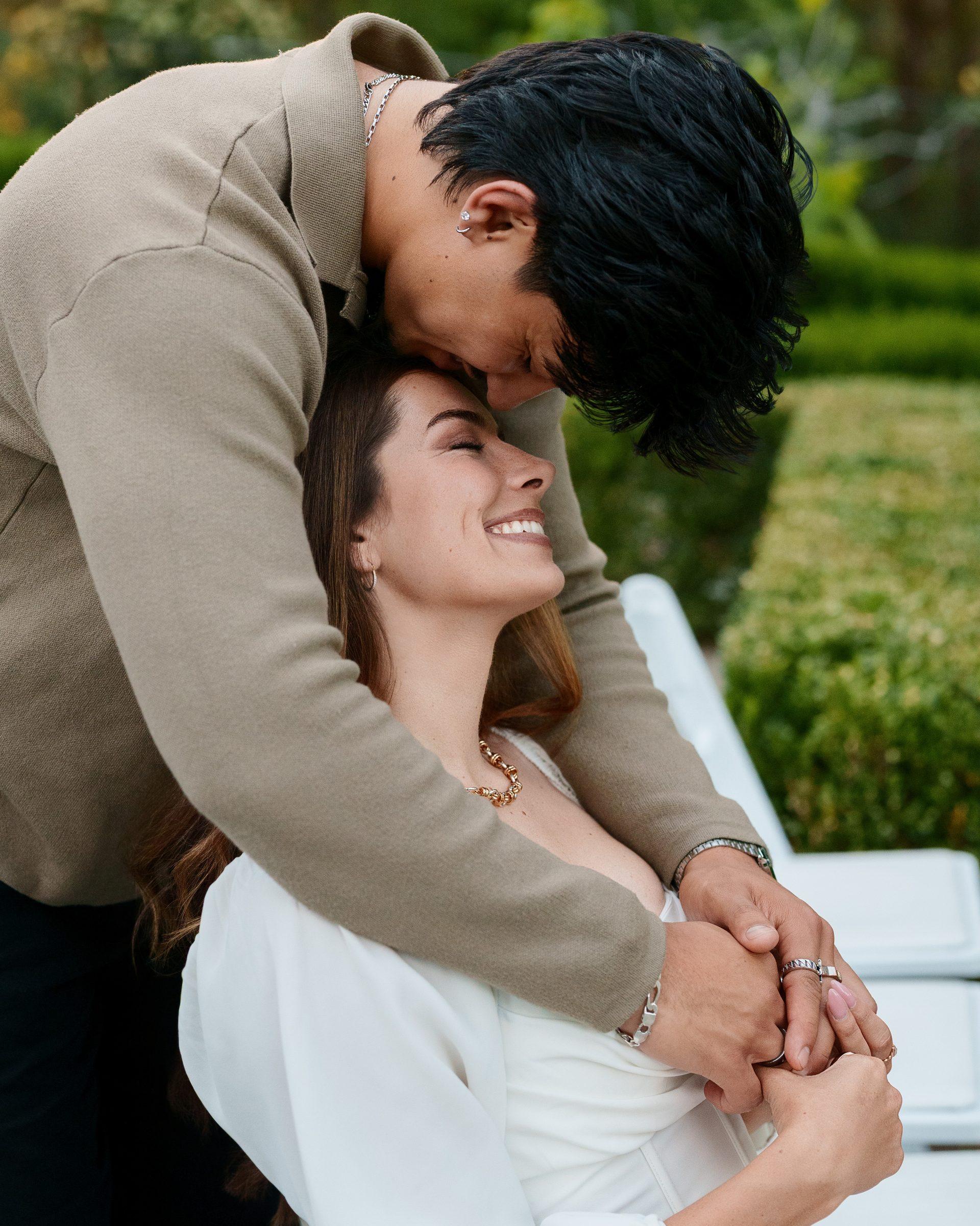 A man in a tan sweater leans down to press his forehead against a woman's temple as she laughs with eyes closed, hands intertwined, lush green hedges and warm garden light behind them at Montalvo Arts Center.