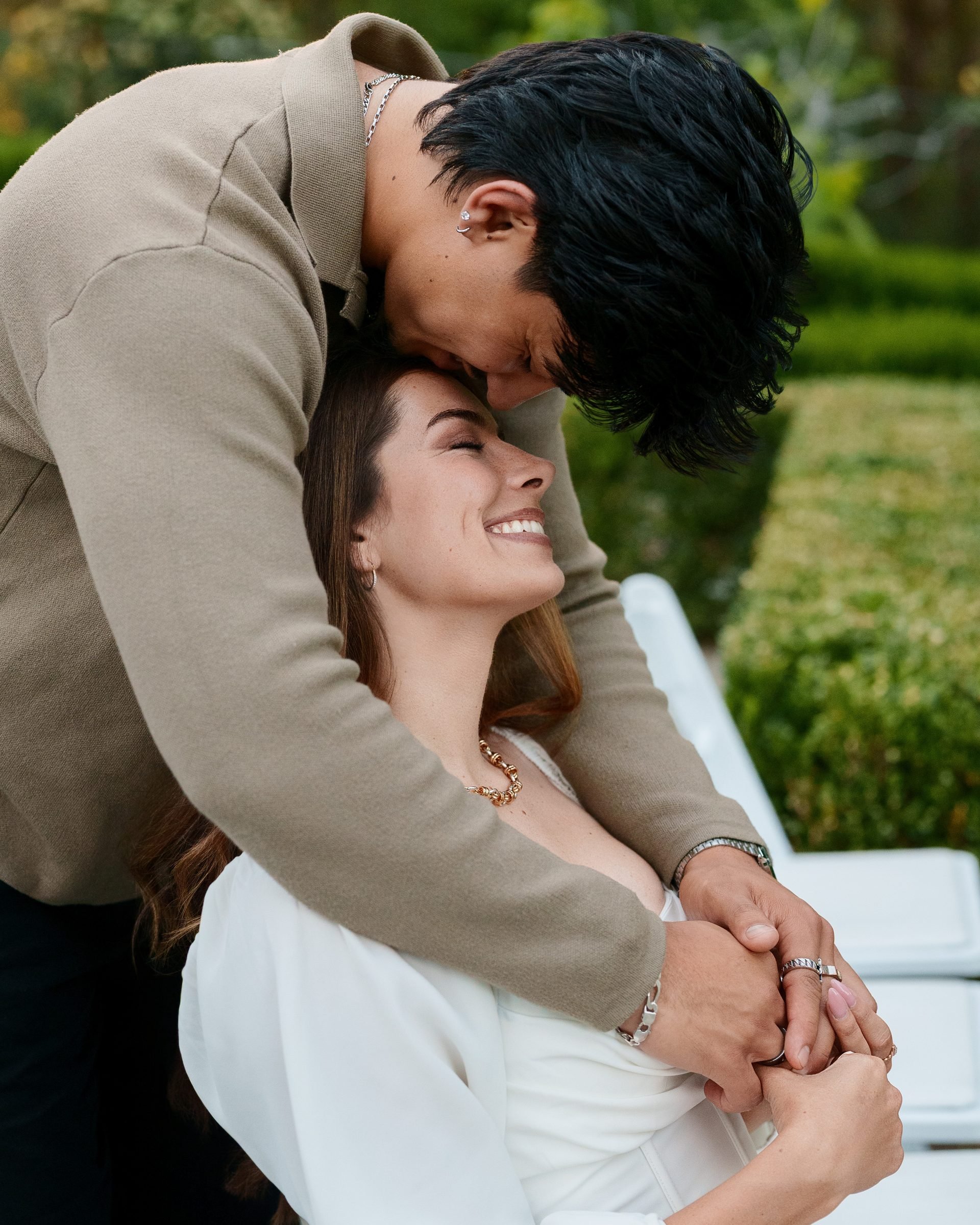 A man in a tan sweater leans down to press his forehead against a woman's temple as she laughs with eyes closed, hands intertwined, lush green hedges and warm garden light behind them at Montalvo Arts Center.