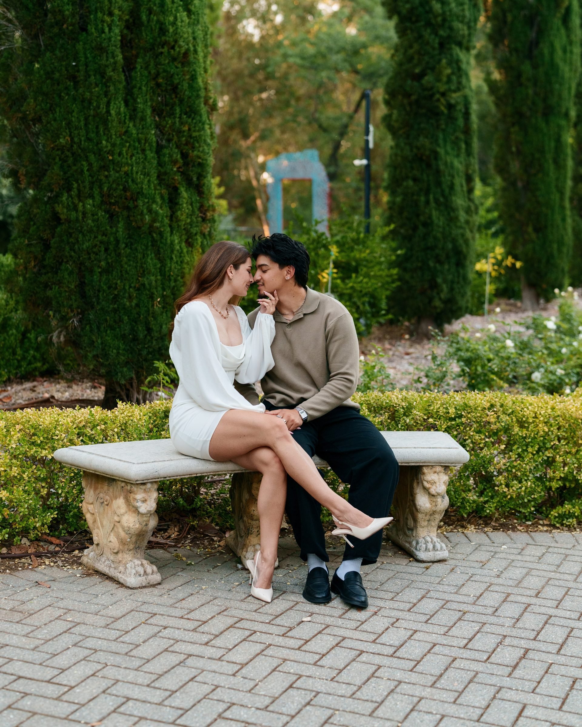 Couple seated close on a stone garden bench at Montalvo Arts Center, faces almost touching in a tender moment, surrounded by Italian cypress trees and manicured hedges in soft golden hour light