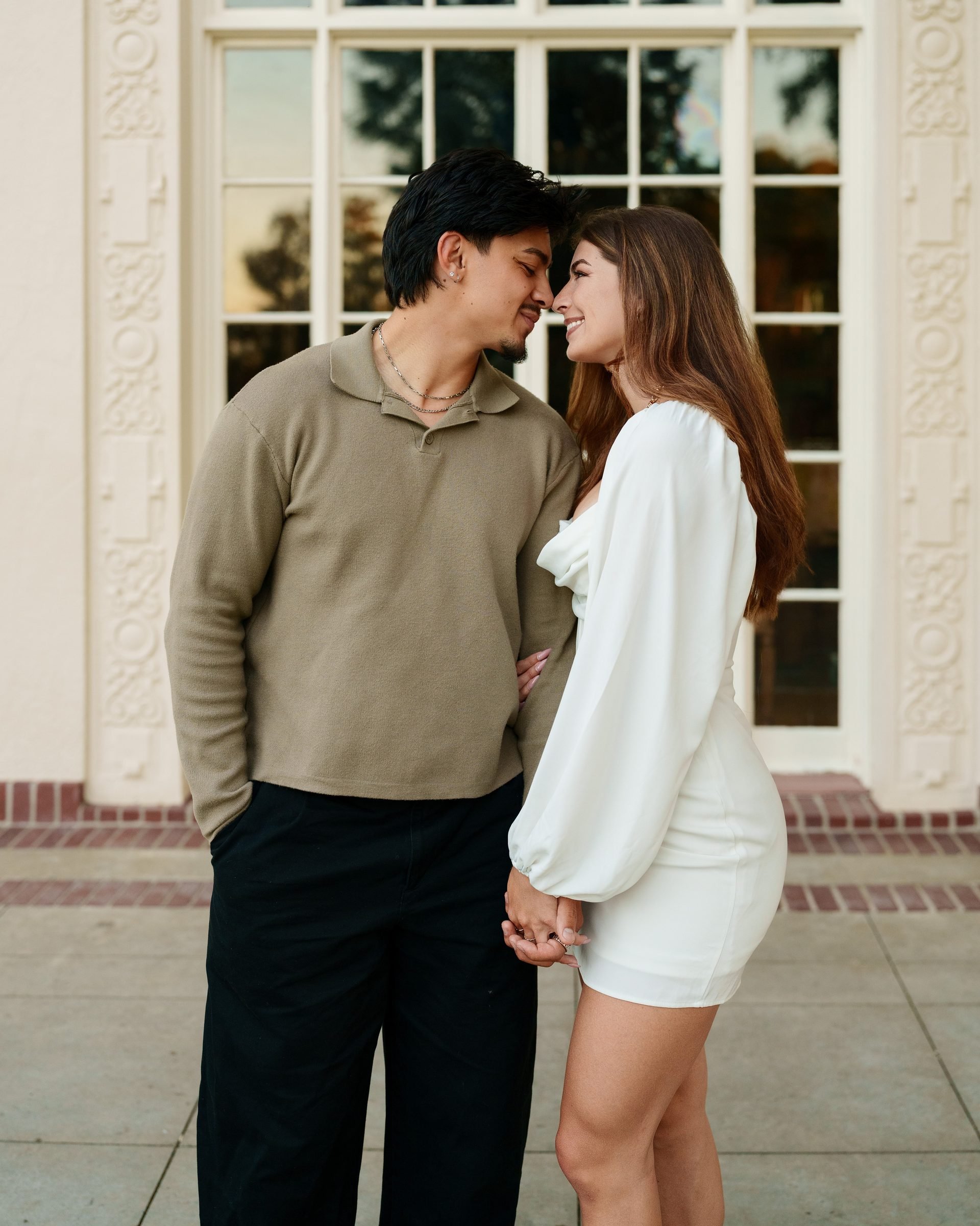 Couple leaning in close, nearly nose-to-nose, in front of an ornate cream-colored building at Montalvo Arts Center during golden hour, she in a white mini dress and he in an olive pullover