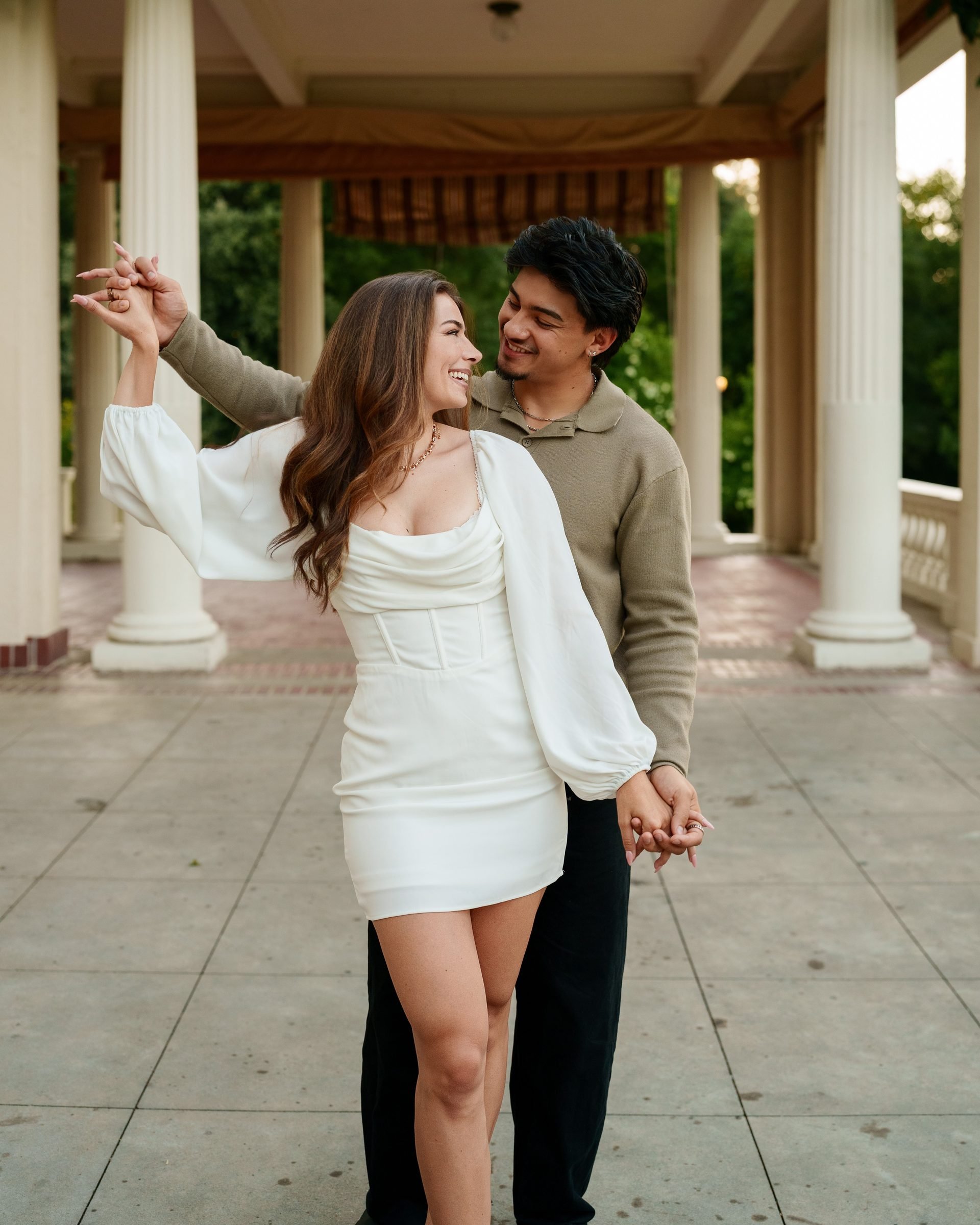 Couple dancing under a columned portico at Montalvo Arts Center during golden hour, she in a white mini dress mid-twirl, he in an olive sweater smiling as he leads her