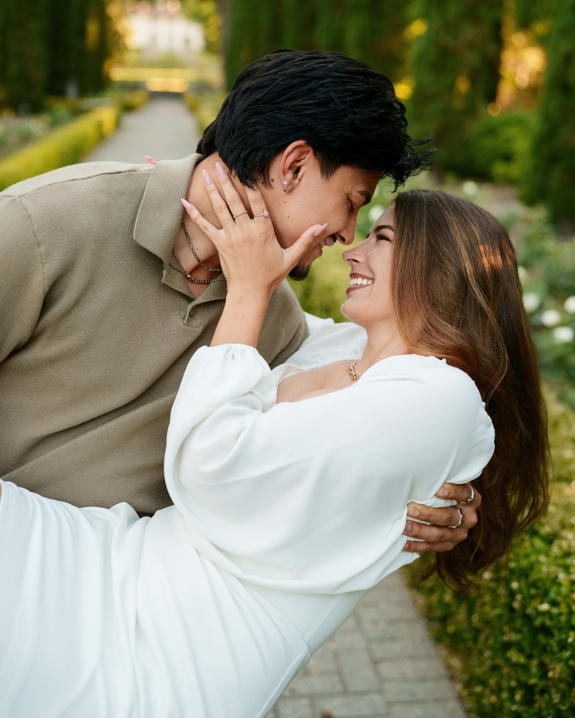Close-up of a couple in a dip pose in a garden, foreheads nearly touching, she laughs up at him with her hand cradling his face, golden hour light warming their skin, he wears a tan shirt, she wears a white mini dress