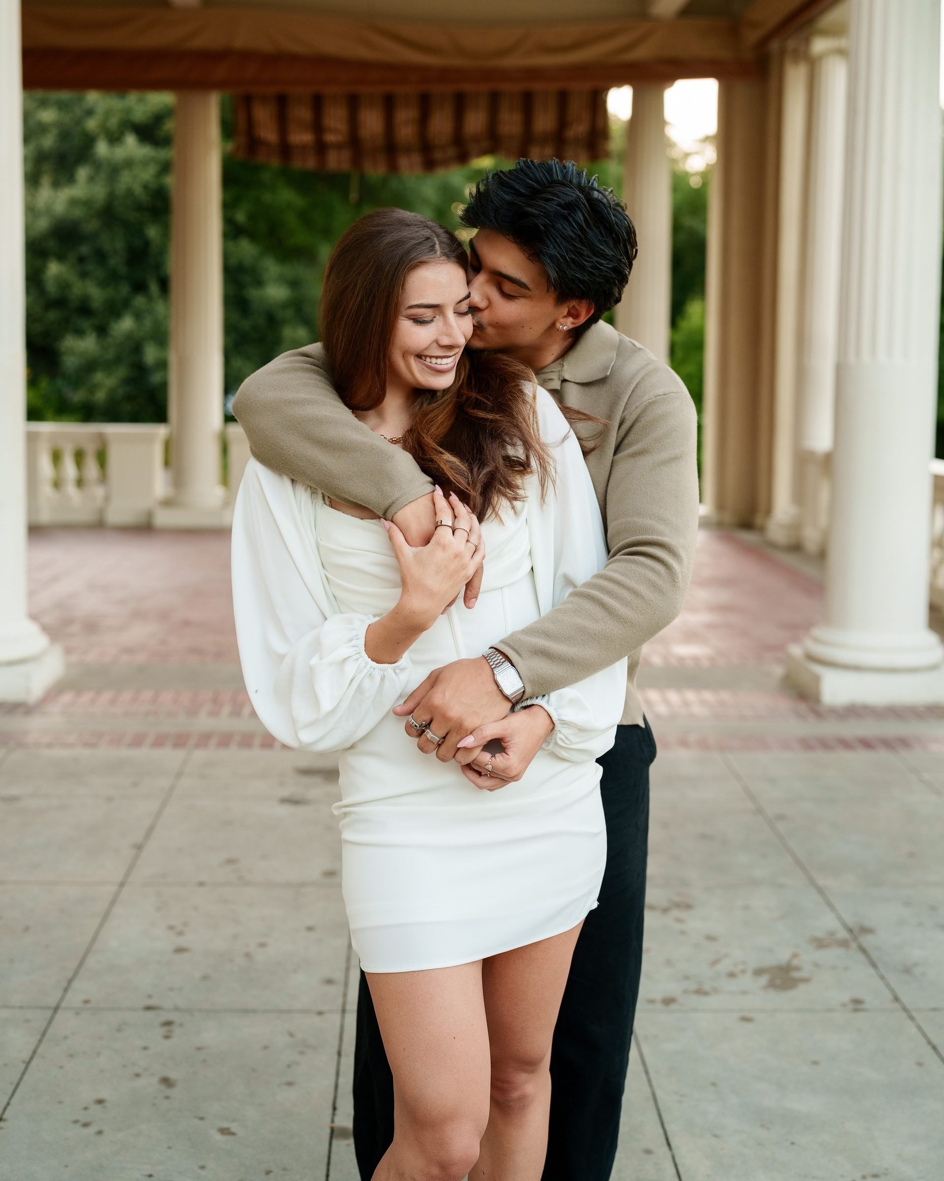 He hugs her from behind and kisses her temple under a classical columned portico at Montalvo Arts Center, she smiles with eyes closed, wearing a white long-sleeve mini dress, he in a tan sweater and black pants.