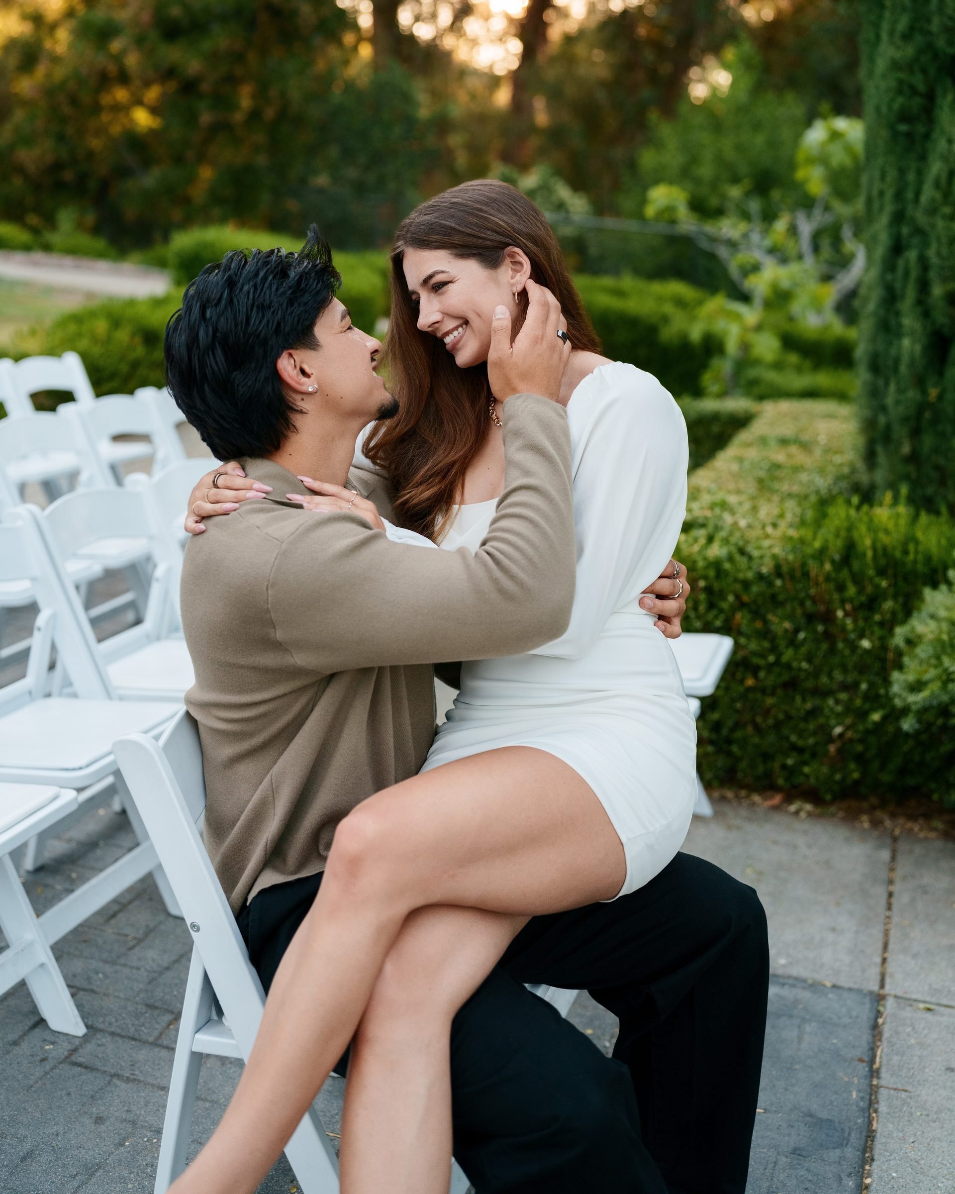 She sits on his lap in a white folding chair amid rows of white chairs at Montalvo Arts Center at golden hour, both leaning in close and smiling, surrounded by lush green hedges and soft warm light.