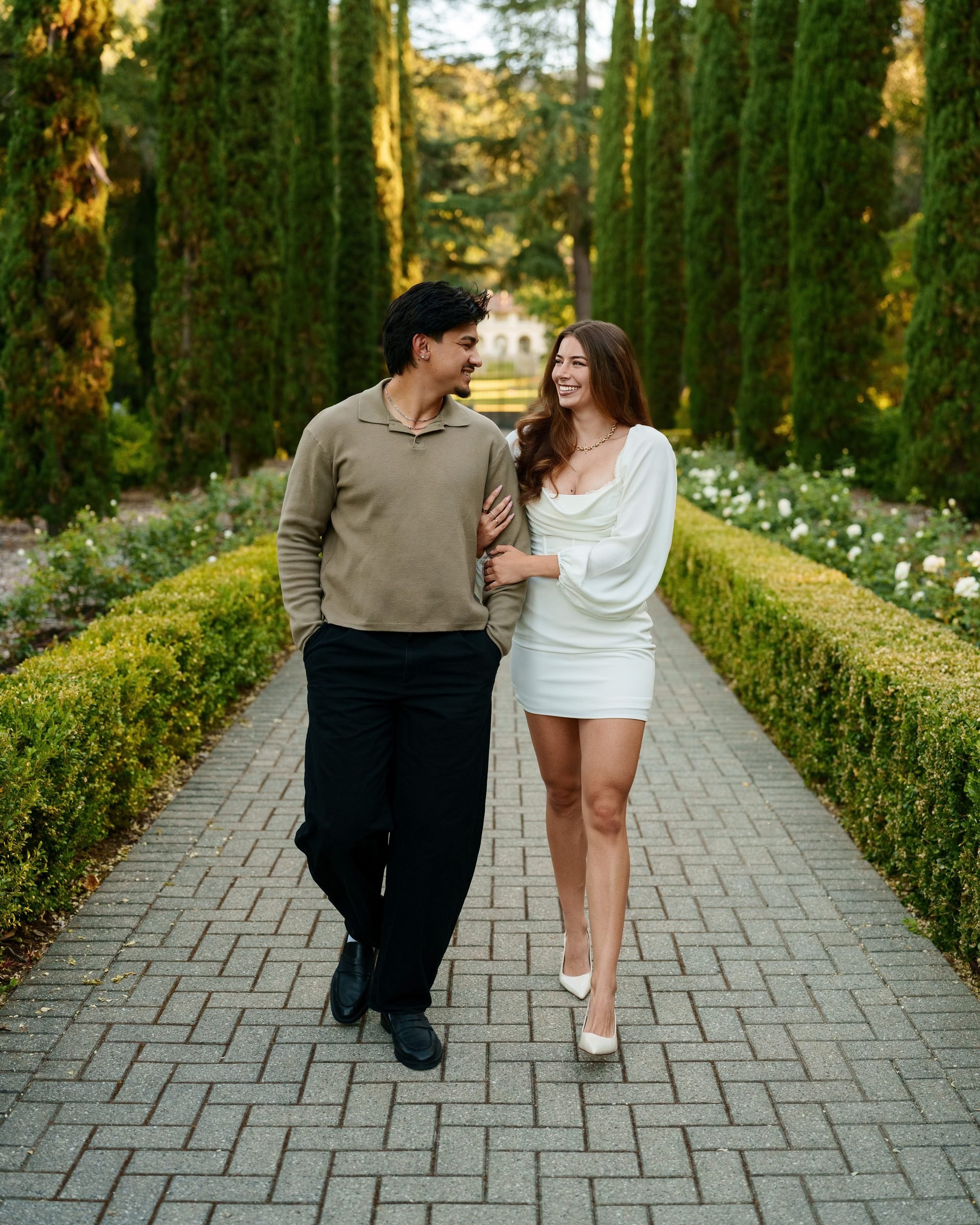 A couple walks arm-in-arm toward the camera along Montalvo's cypress-lined brick path at golden hour, laughing together. She wears a white cowl-neck mini dress with white heels; he wears a tan knit sweater and black trousers.