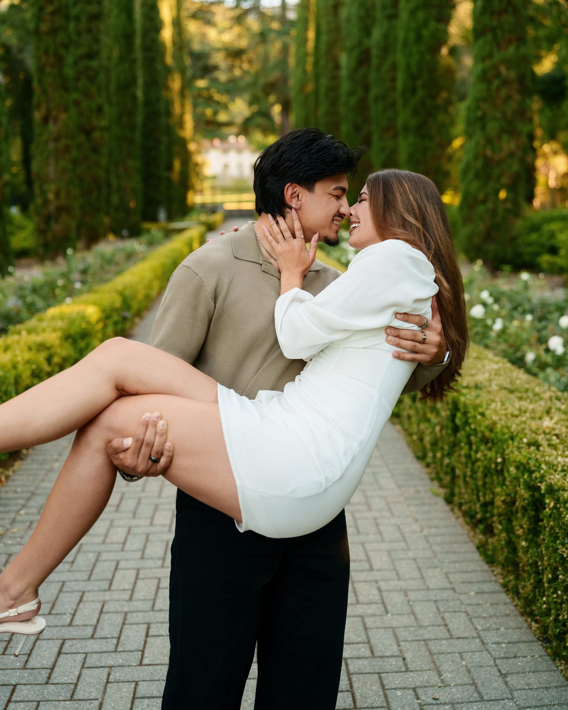 Man lifts woman off the ground on a brick garden path at Montalvo Arts Center, both laughing and leaning noses together, she holds his face in her hands, golden hour light glows behind them through cypress trees