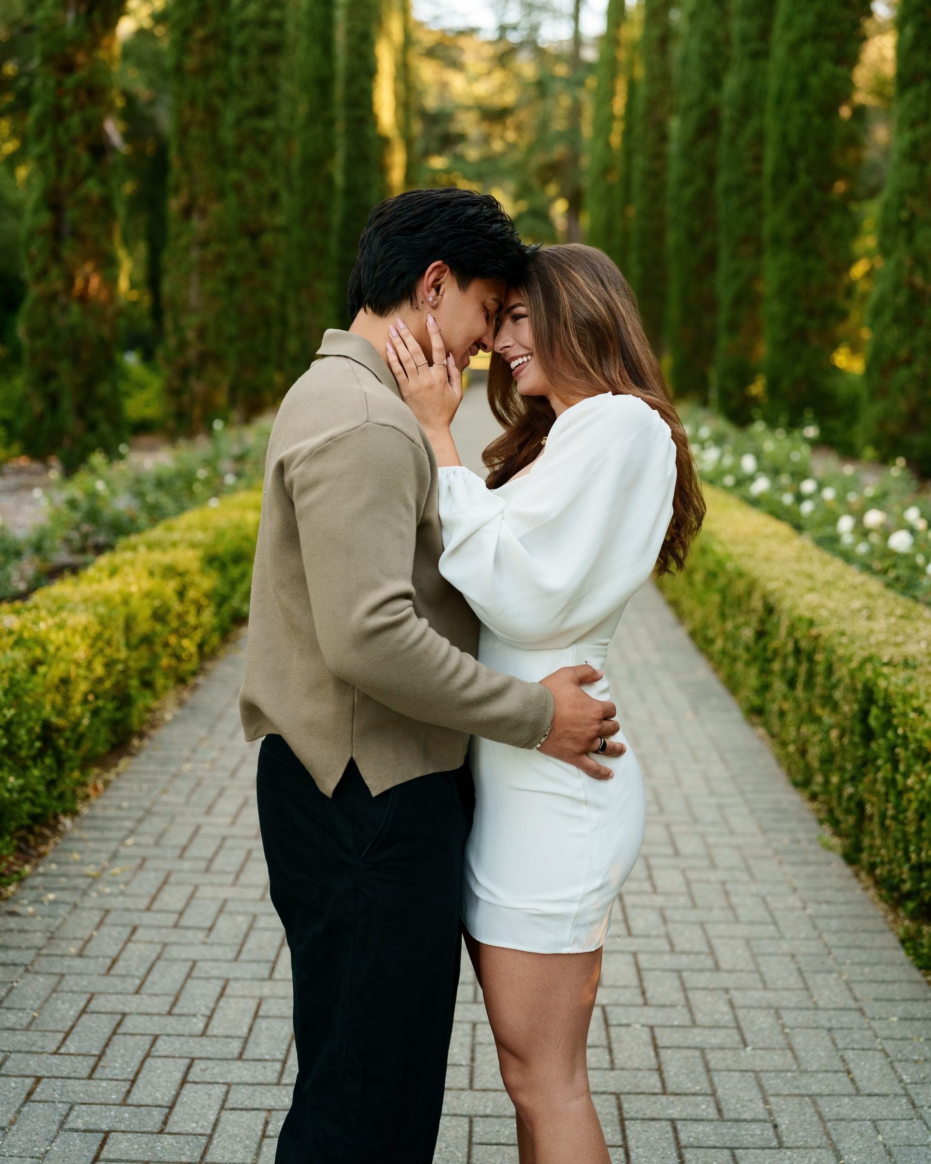Couple standing forehead-to-forehead on a symmetrical brick garden path lined with tall Italian cypresses at golden hour, she smiles broadly, he leans in close, both in neutral tones