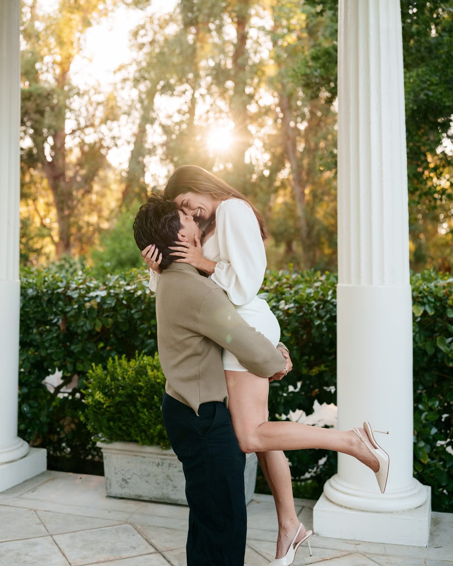 Man lifts woman off the ground between white columns at Montalvo Arts Center during golden hour; she laughs with head tilted back, wearing a white mini dress and slingback heels while he holds her in a tan knit sweater and black trousers.