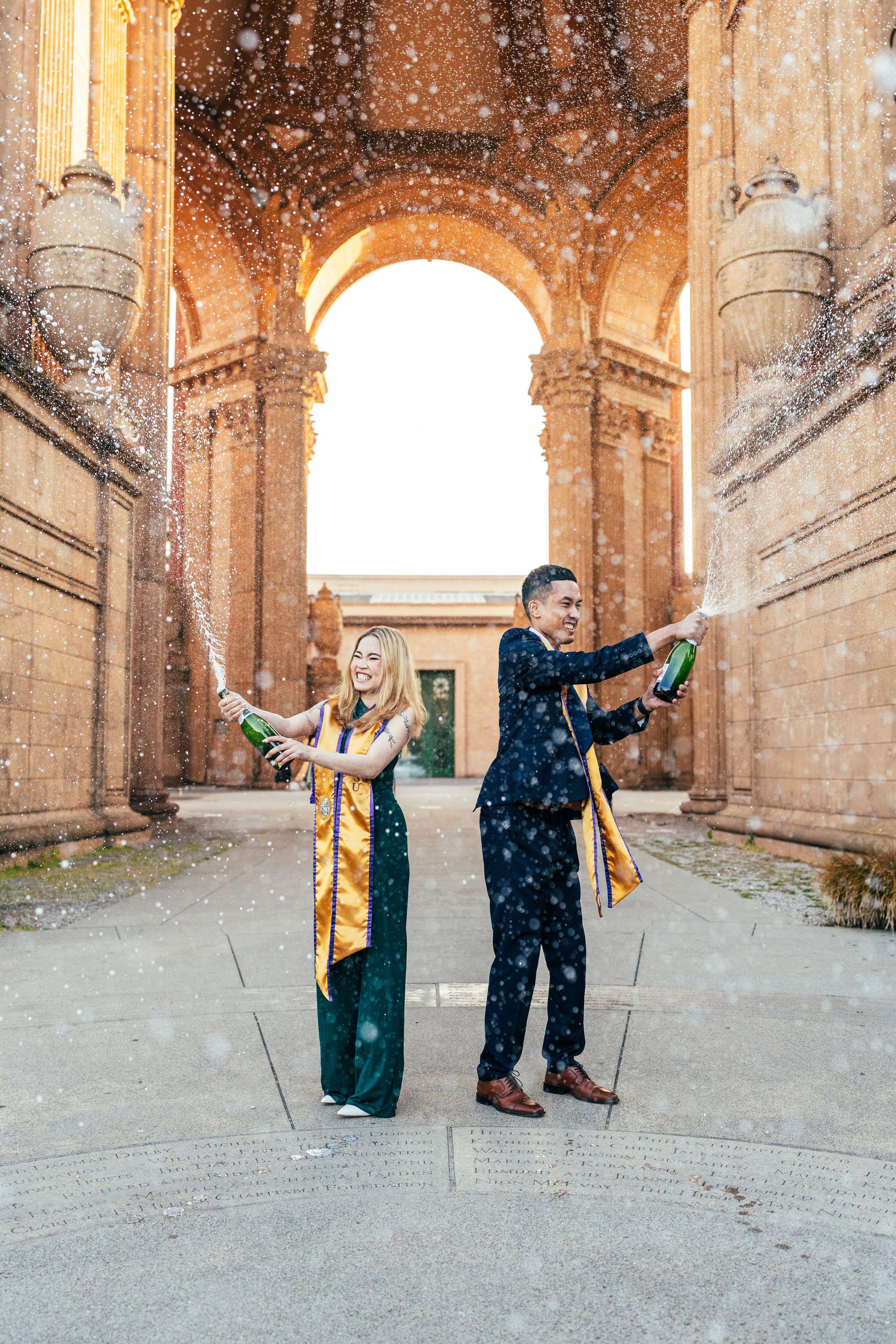 Two graduates in gold sashes spray champagne under the Palace of Fine Arts rotunda at golden hour