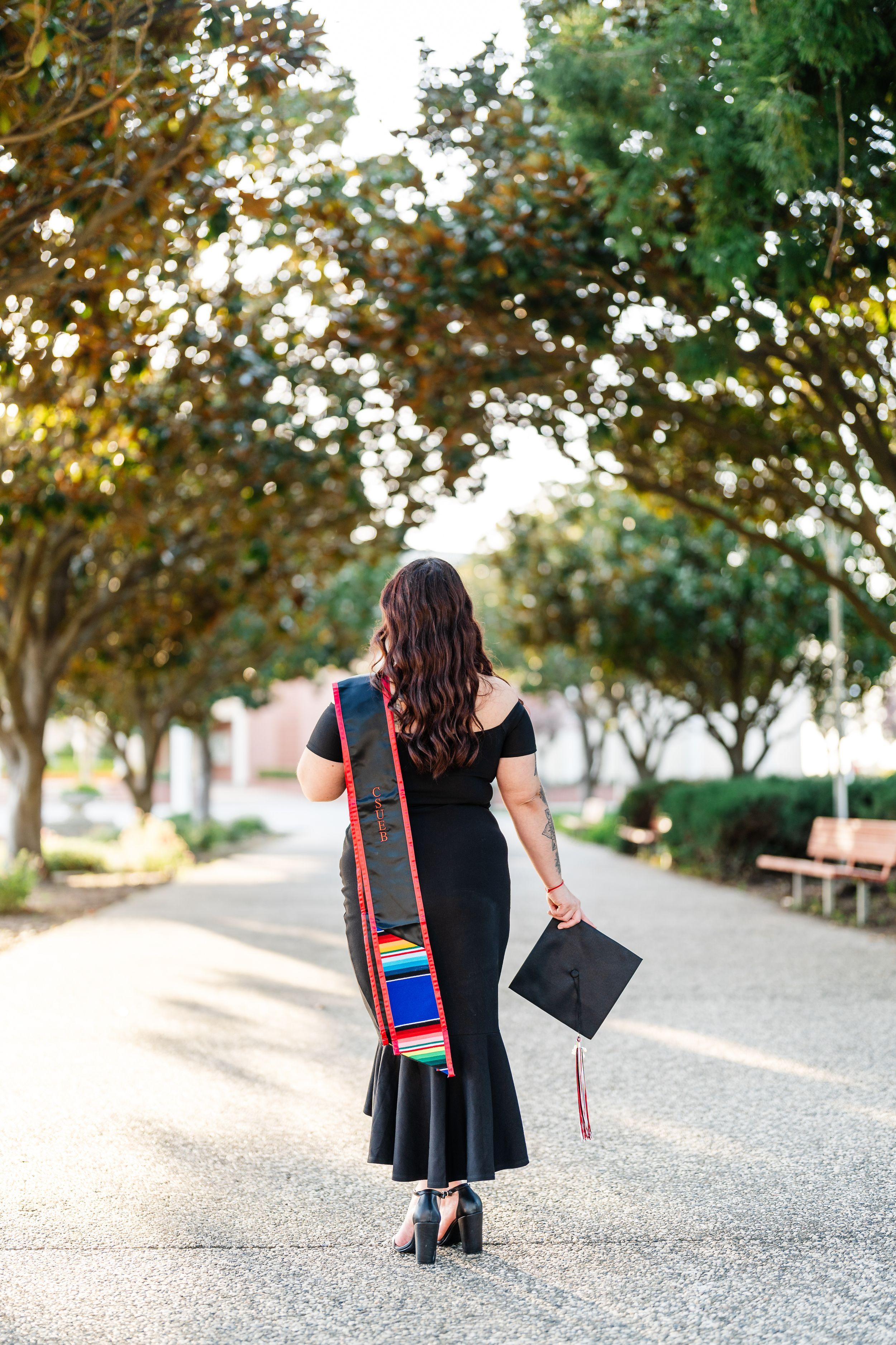 Bay Area graduation portrait of a CSUEB graduate in a black dress holding her cap, serape stole draped, on a tree-lined path.