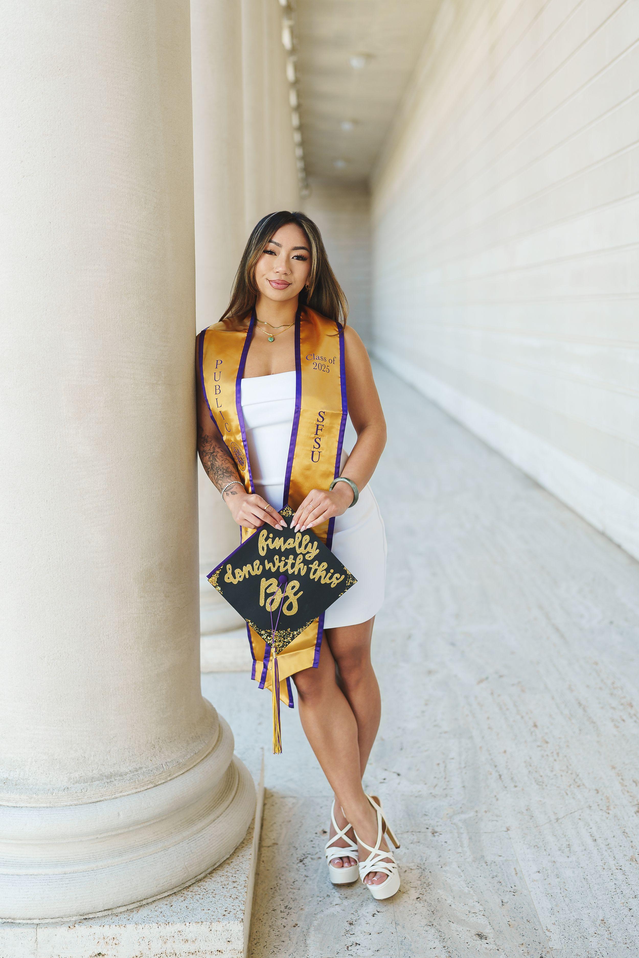 Bay Area graduation portrait of SFSU grad in white dress and gold sash leaning on column, holding decorated cap.