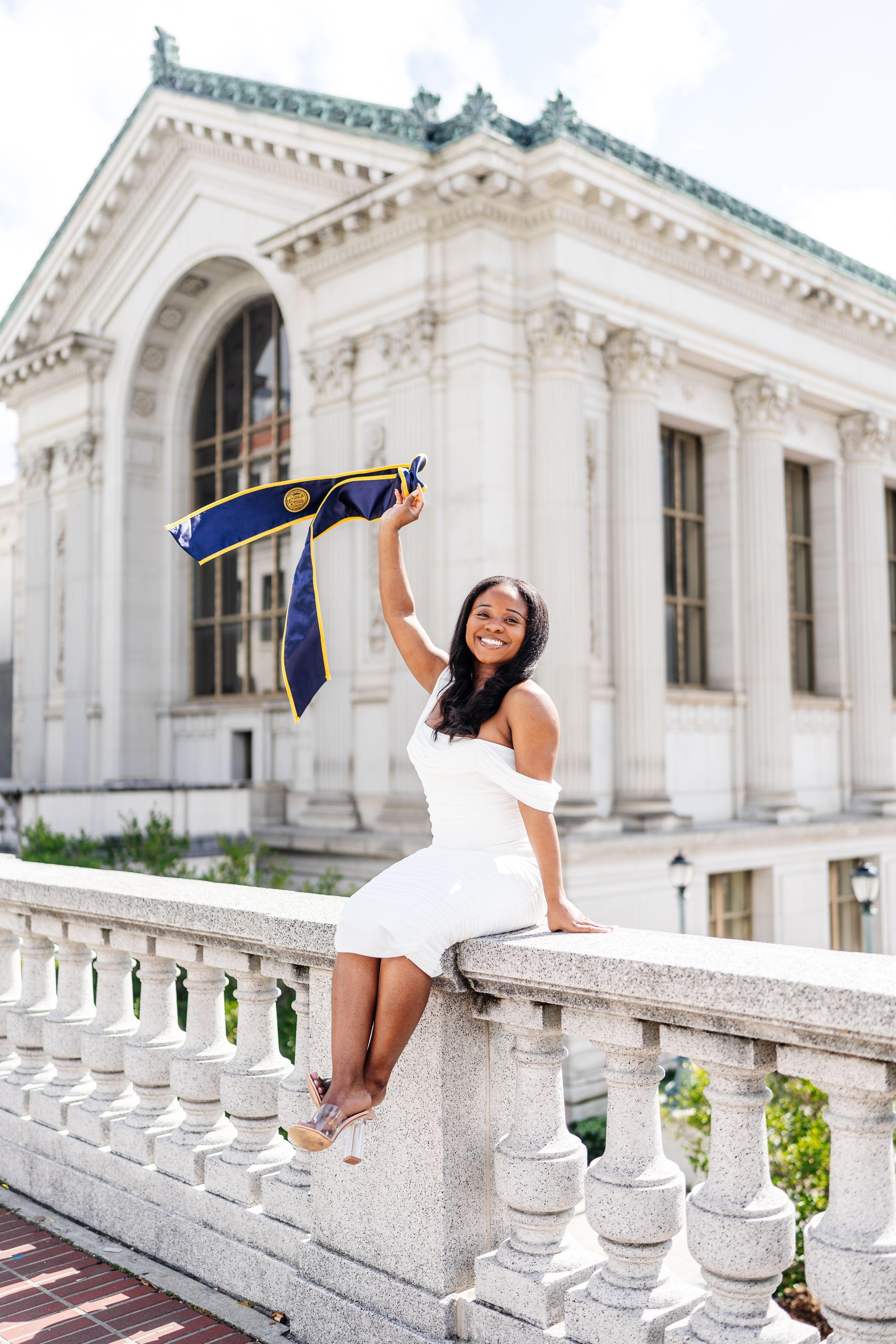UC Berkeley graduate in a white dress sits on a stone balustrade waving a navy and gold stole near Doe Library.