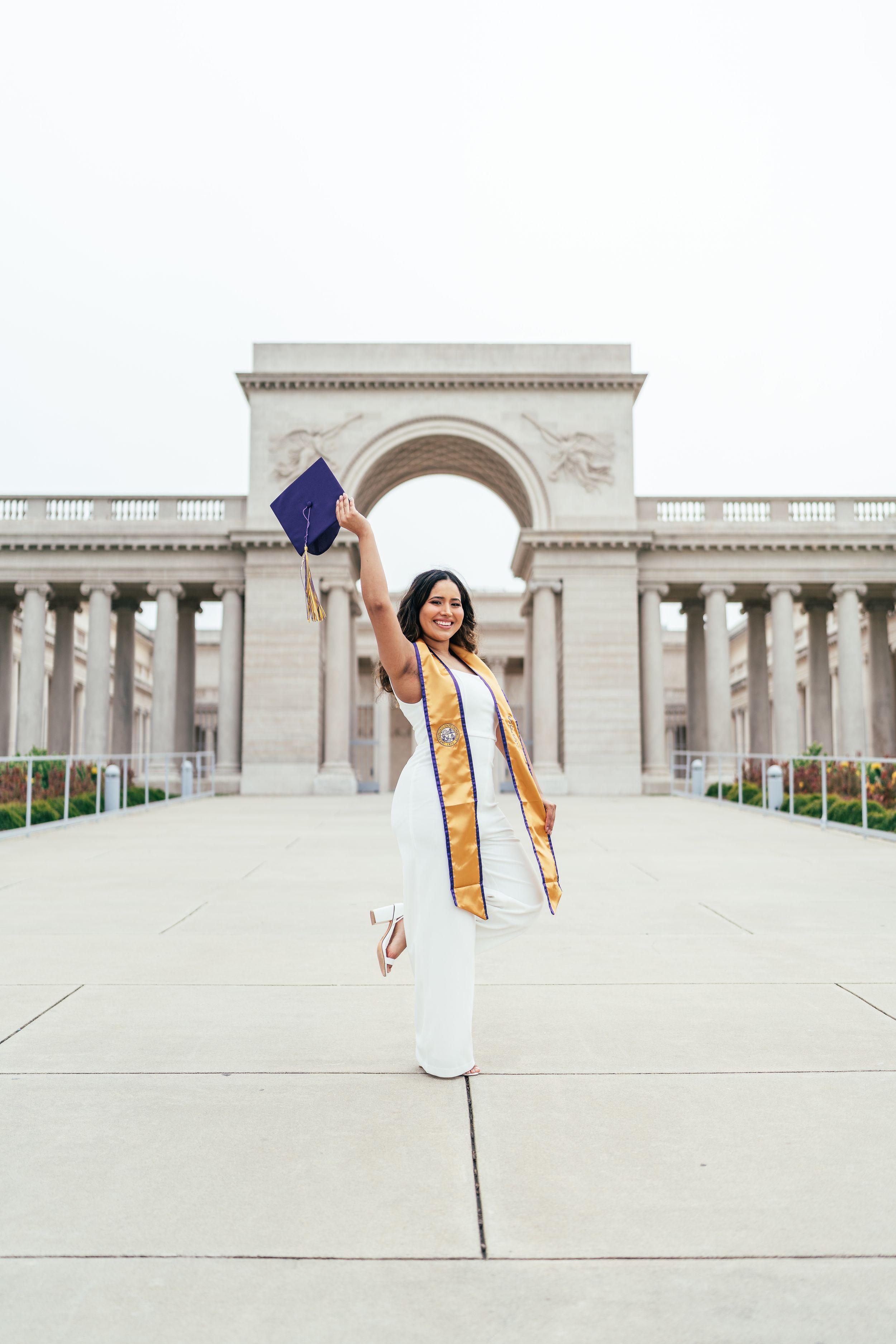 Bay Area graduation portrait of a graduate in a white dress and gold sash raising her purple cap at the Legion of Honor.