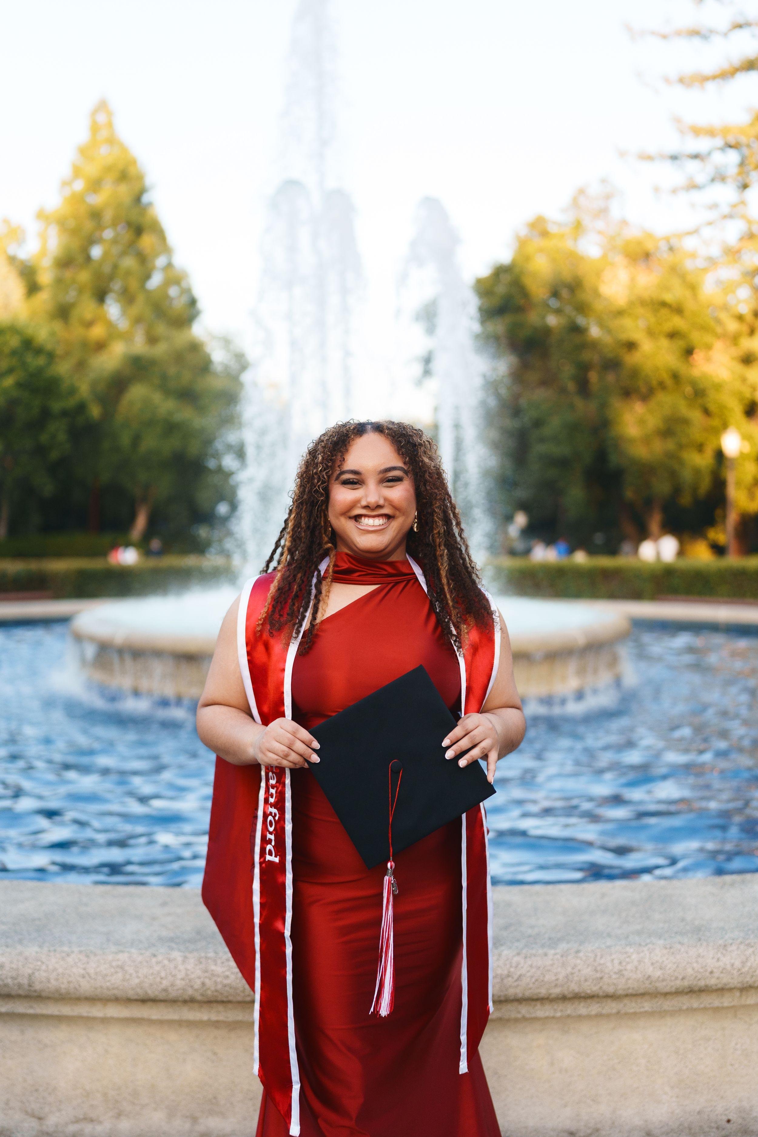 Smiling Stanford graduate in red dress and sash holds black cap by a fountain at golden hour, Bay Area graduation portrait.