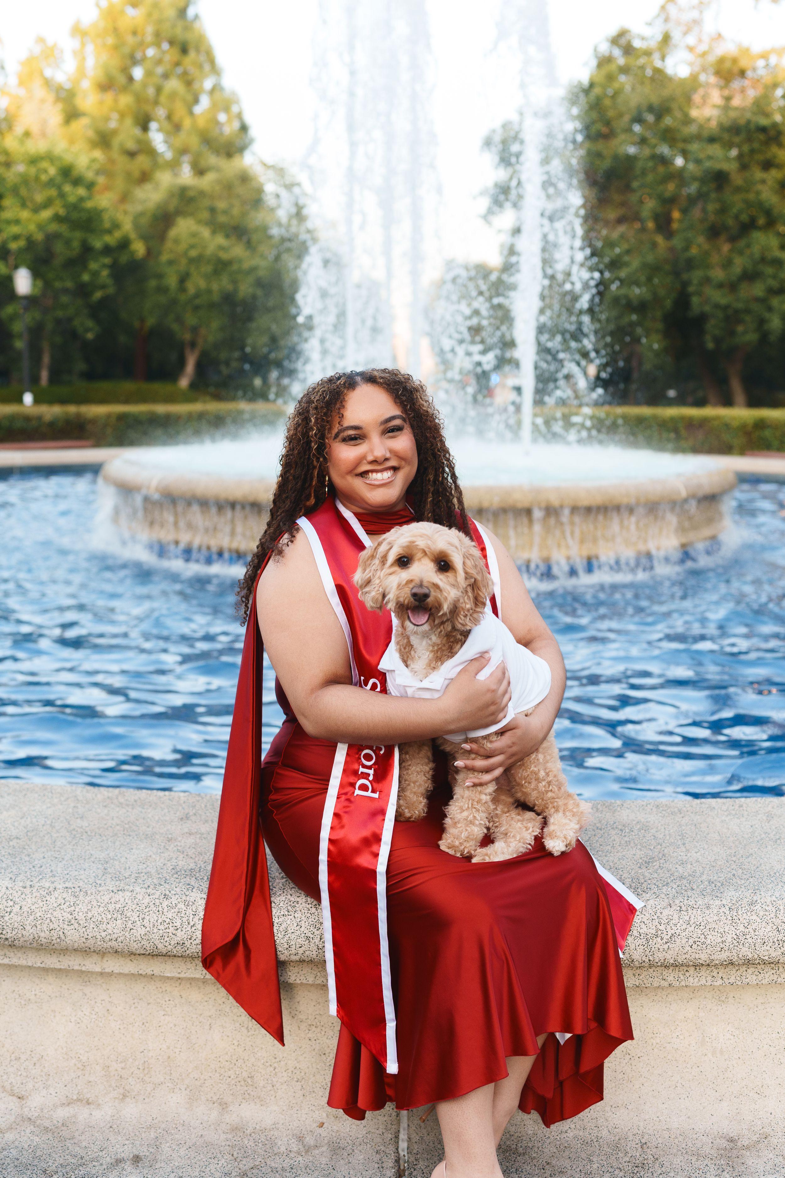 Bay Area graduation portrait of a smiling Stanford grad in a red satin dress and sash holding her small dog by a fountain.