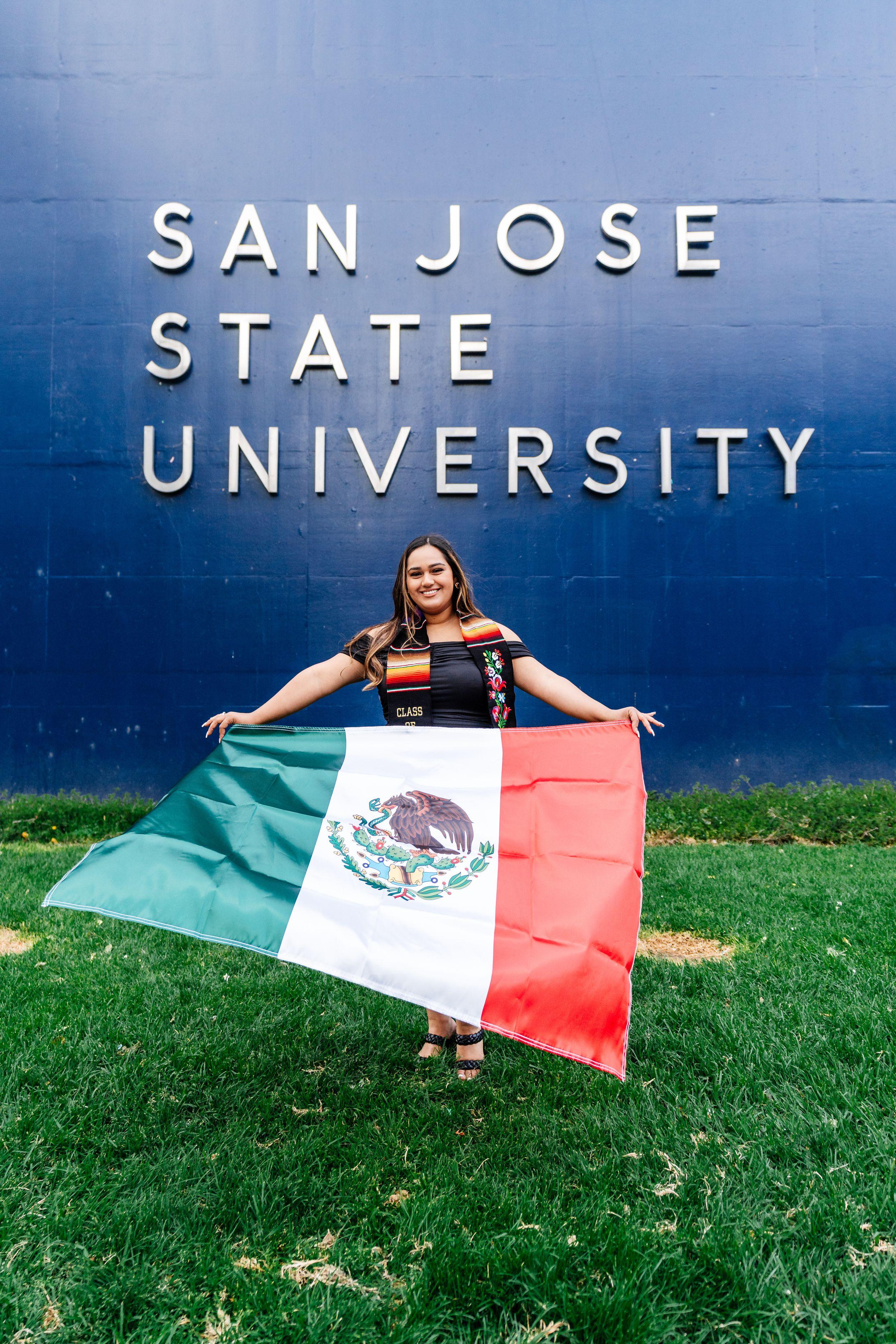 Bay Area graduation portrait of a smiling SJSU graduate in a black dress and serape stole holding a Mexican flag on the lawn