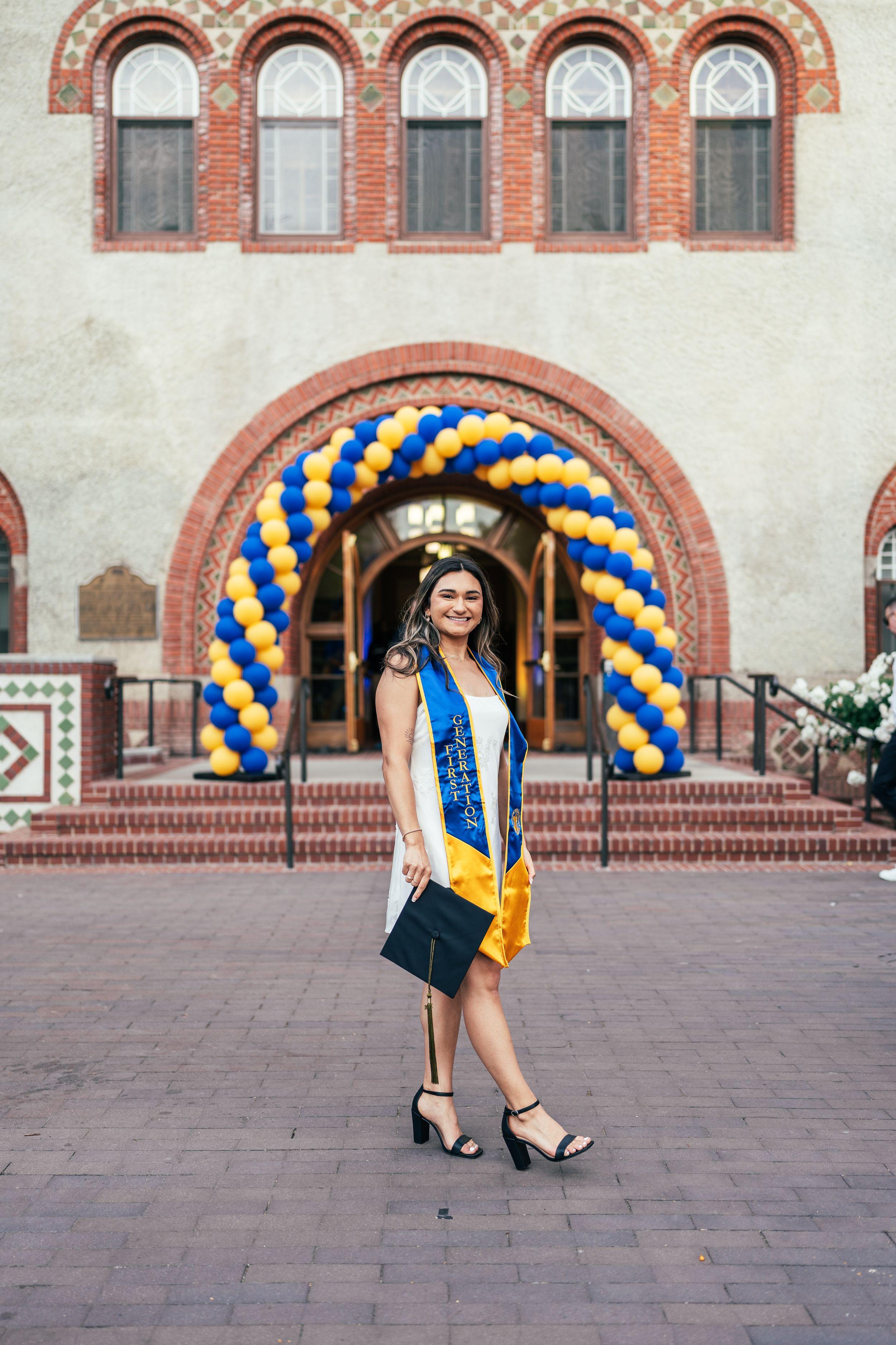 Bay Area graduation portrait of a smiling woman in a white dress and blue First Generation stole holding her cap beneath a