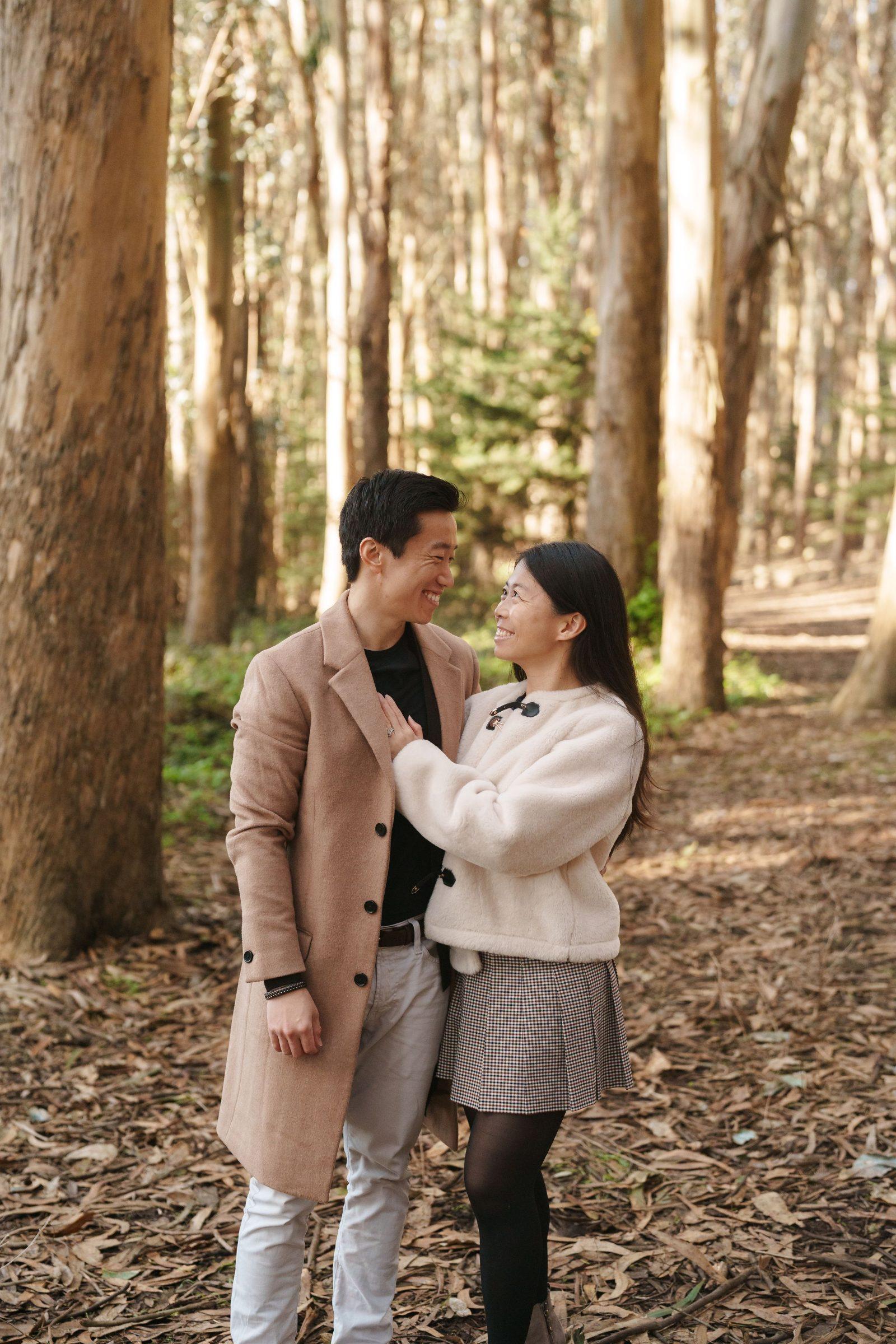Bay Area family portrait of a couple laughing together among eucalyptus trees, wearing camel coat and cream jacket in warm